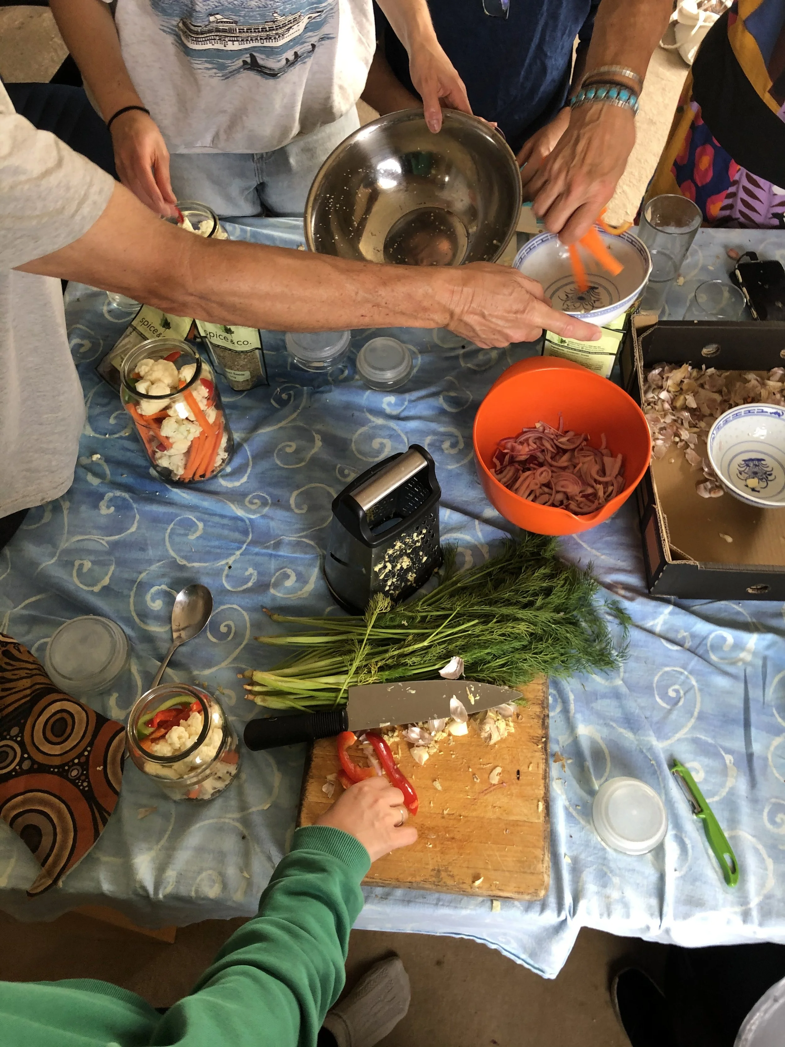 People preparing food together around a table with chopping, mixing, and ingredients like dill, garlic, onions, and cauliflower.
