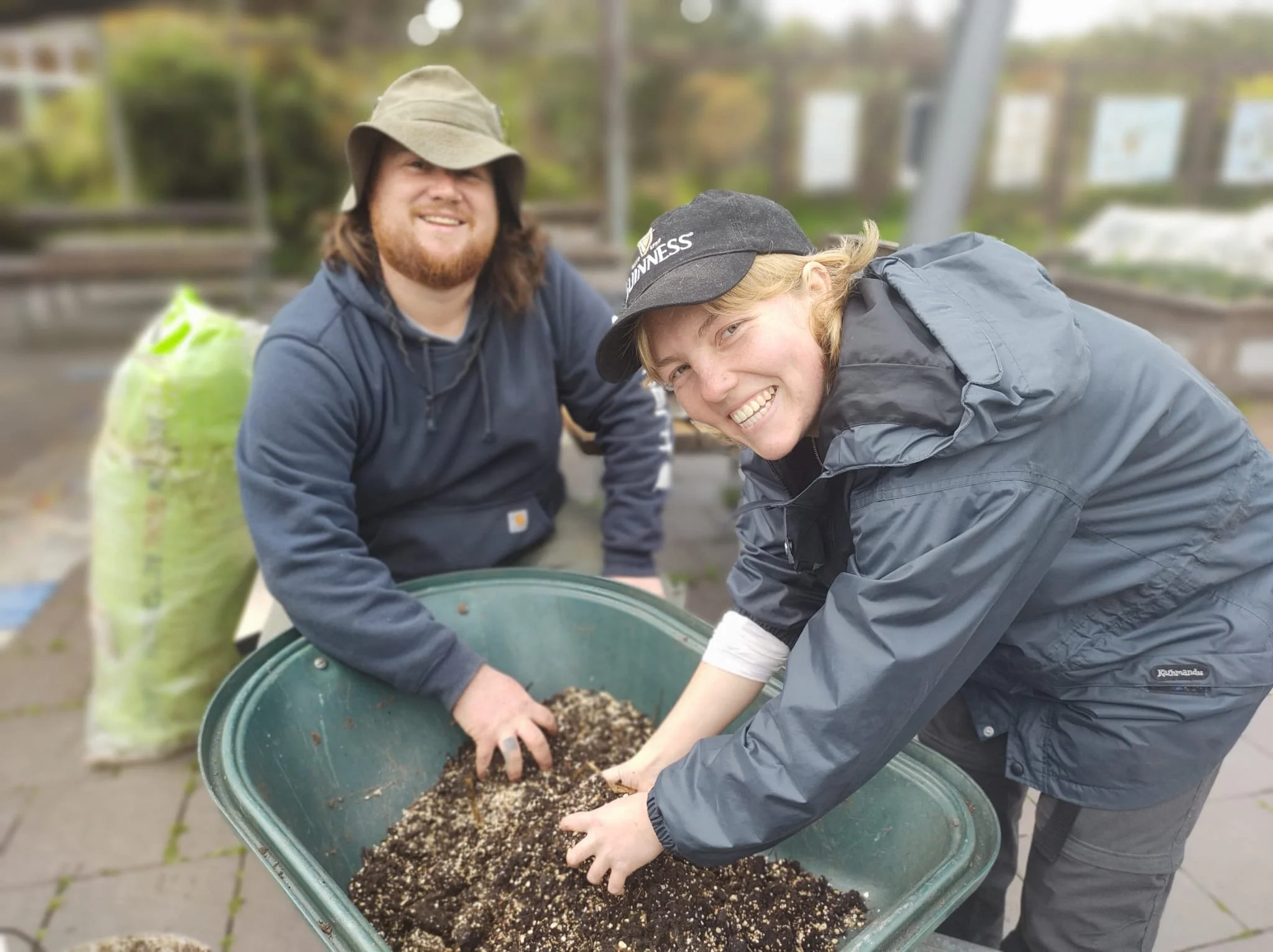 Two men gardening outdoors, one wearing a wide-brimmed hat and hoodie, the other wearing a baseball cap and jacket, smiling while handling soil in a wheelbarrow.