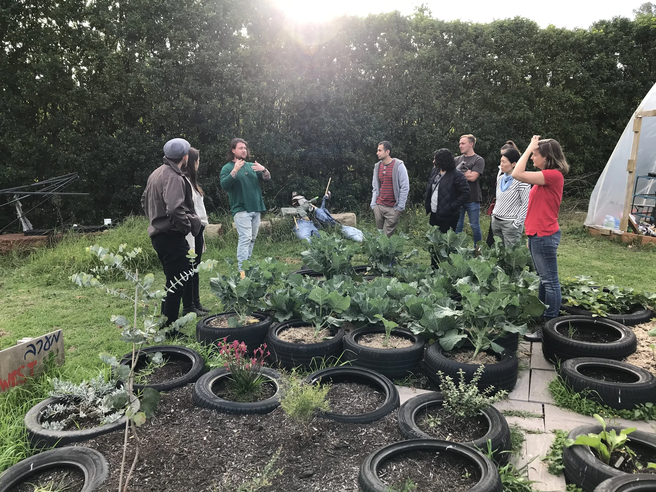 A group of people gathered outdoors near a garden with plants growing in repurposed tires arranged in rows. Someone appears to be giving a presentation or leading a discussion during daylight with trees in the background.