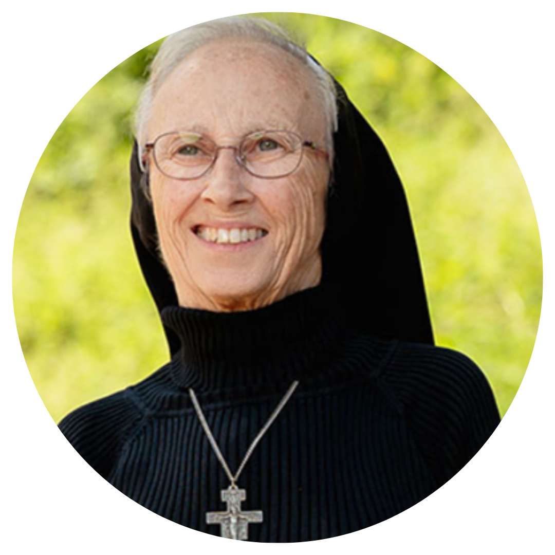 Close-up of a smiling elderly woman wearing glasses, a black habit, and a cross necklace, outdoors with green foliage in the background.