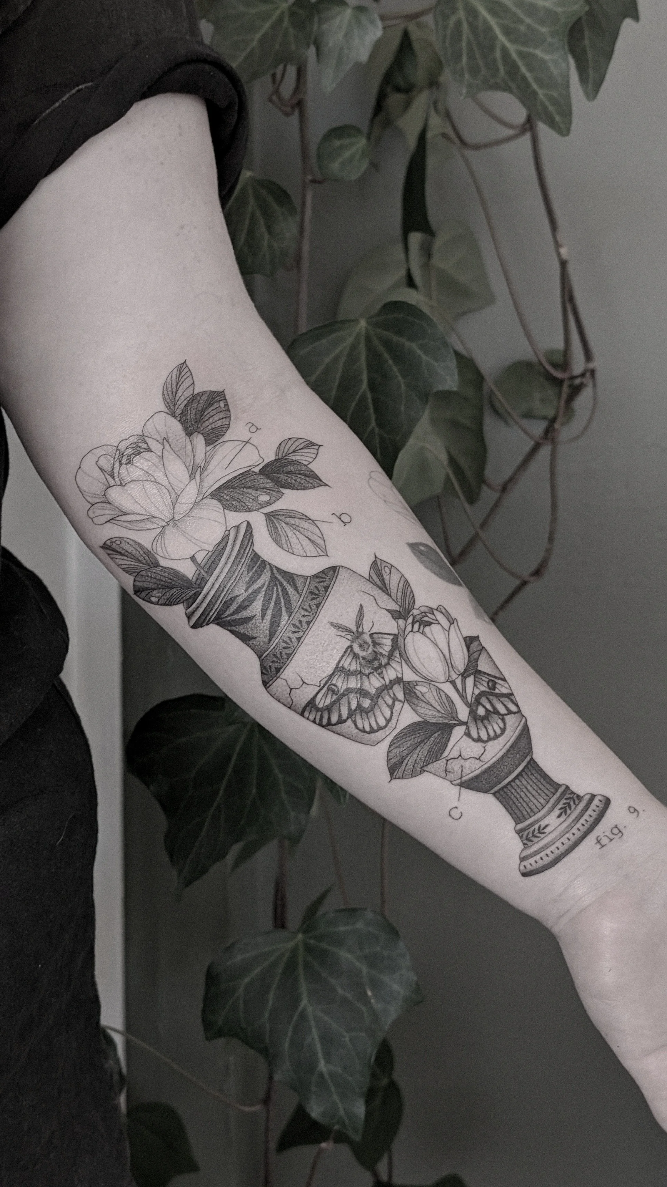 A black and gray tattoo of a broken vase with a moth with peonies coming out, featuring detailed line work, on a person's forearm against a background of green ivy leaves.