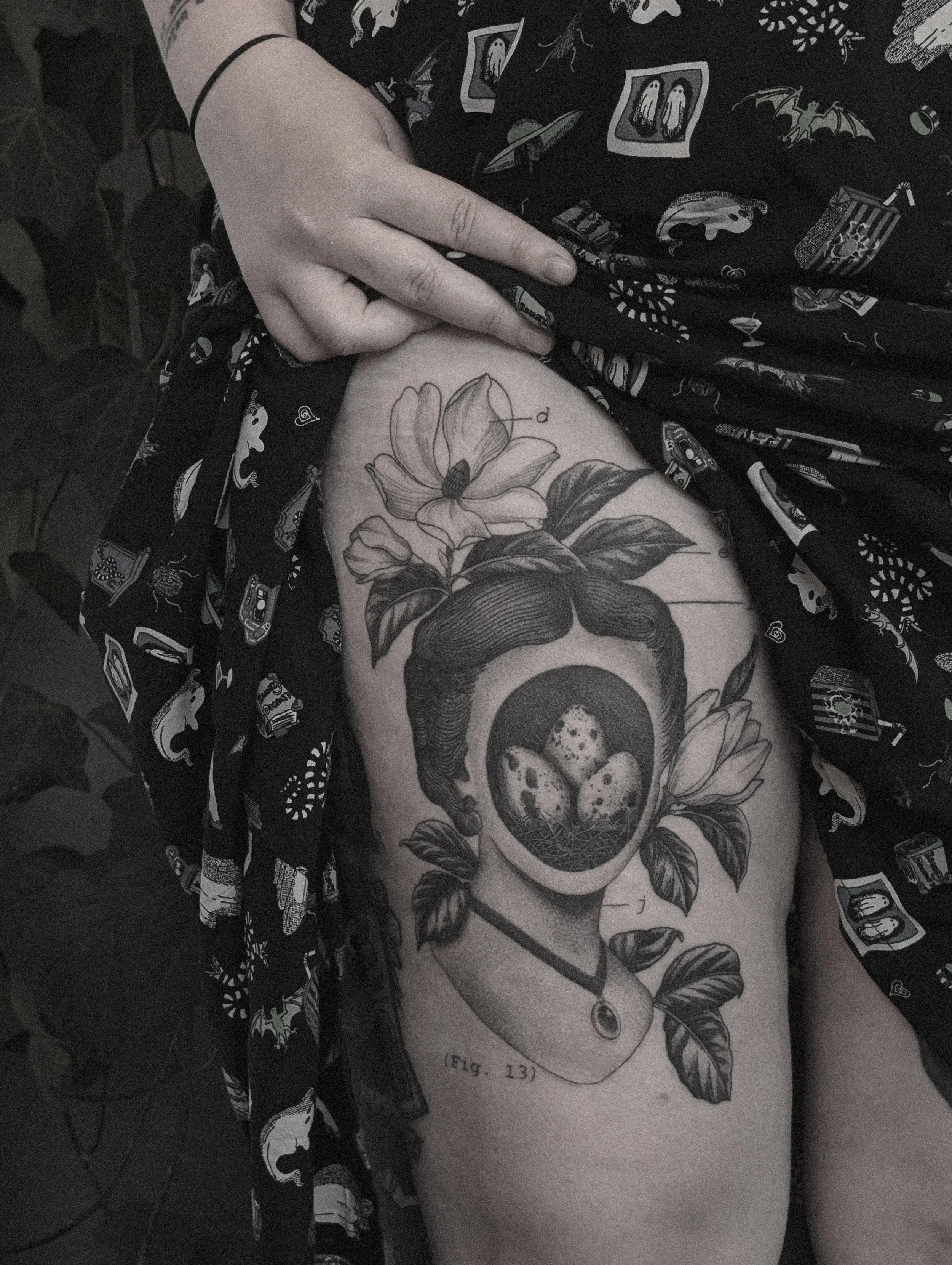 Tattoo on person's upper thigh featuring a woman's head with floral elements, a nest with eggs inside the head, and a necklace with a gemstone, with a caption '(Fig. 13)'.