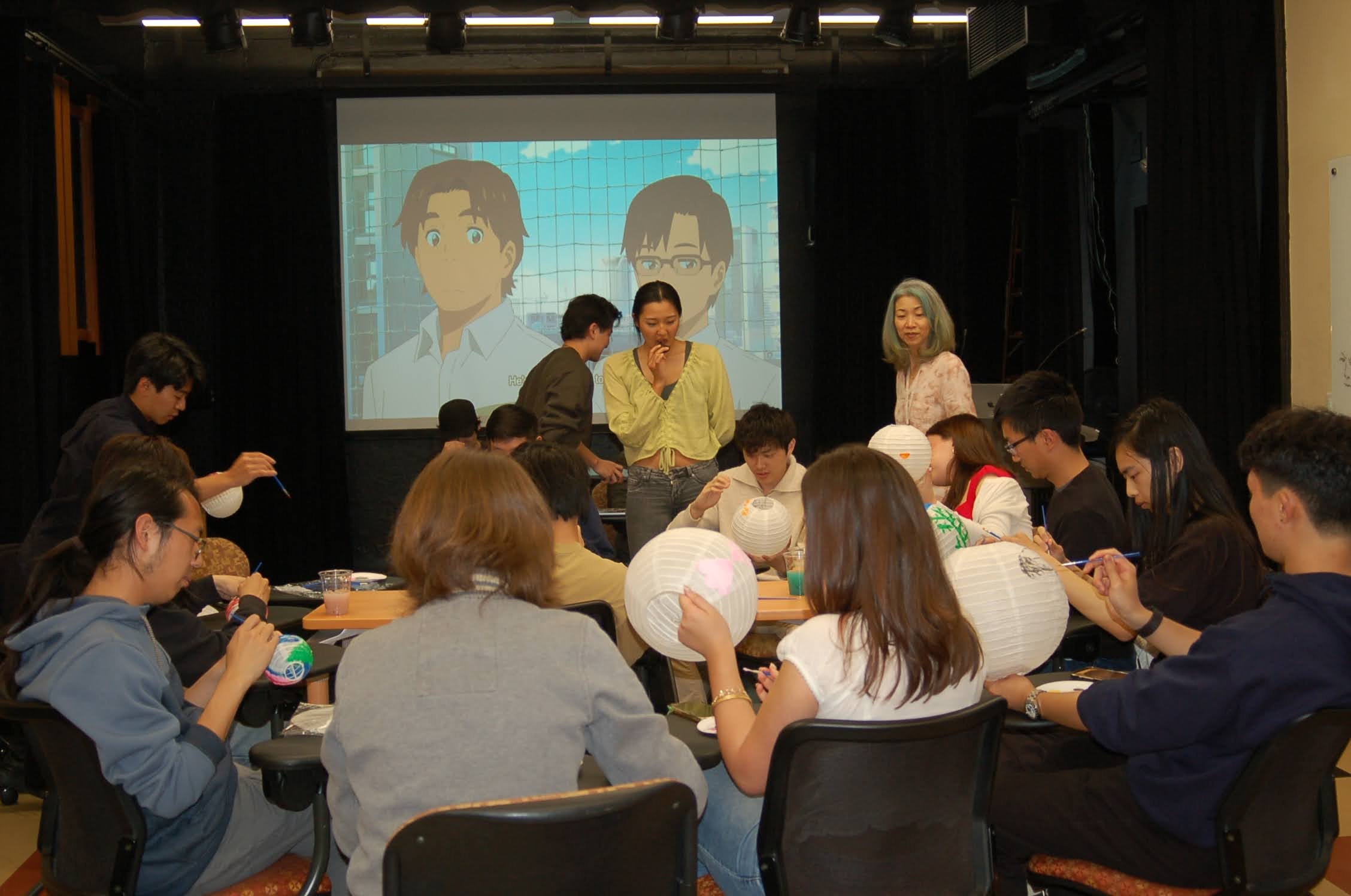 Group of people sitting around tables, painting paper lanterns, with a large screen displaying animated characters in the background.