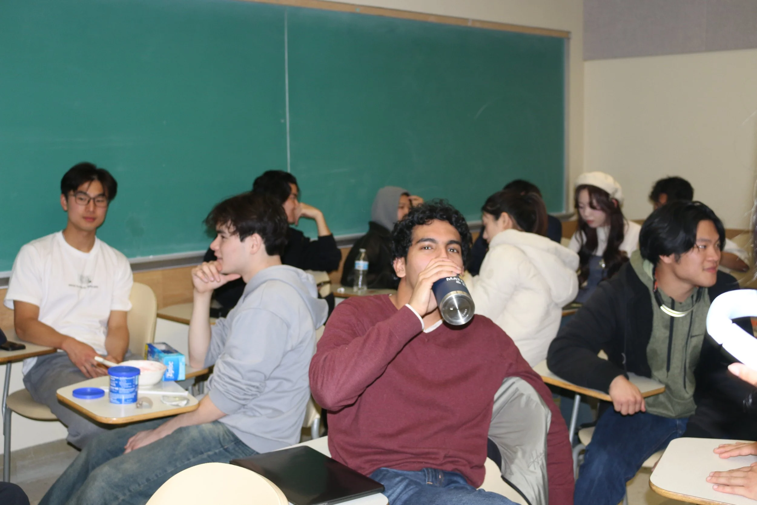 Students in a classroom, some sitting at desks, one drinking from a tumbler, and others engaging in various activities.