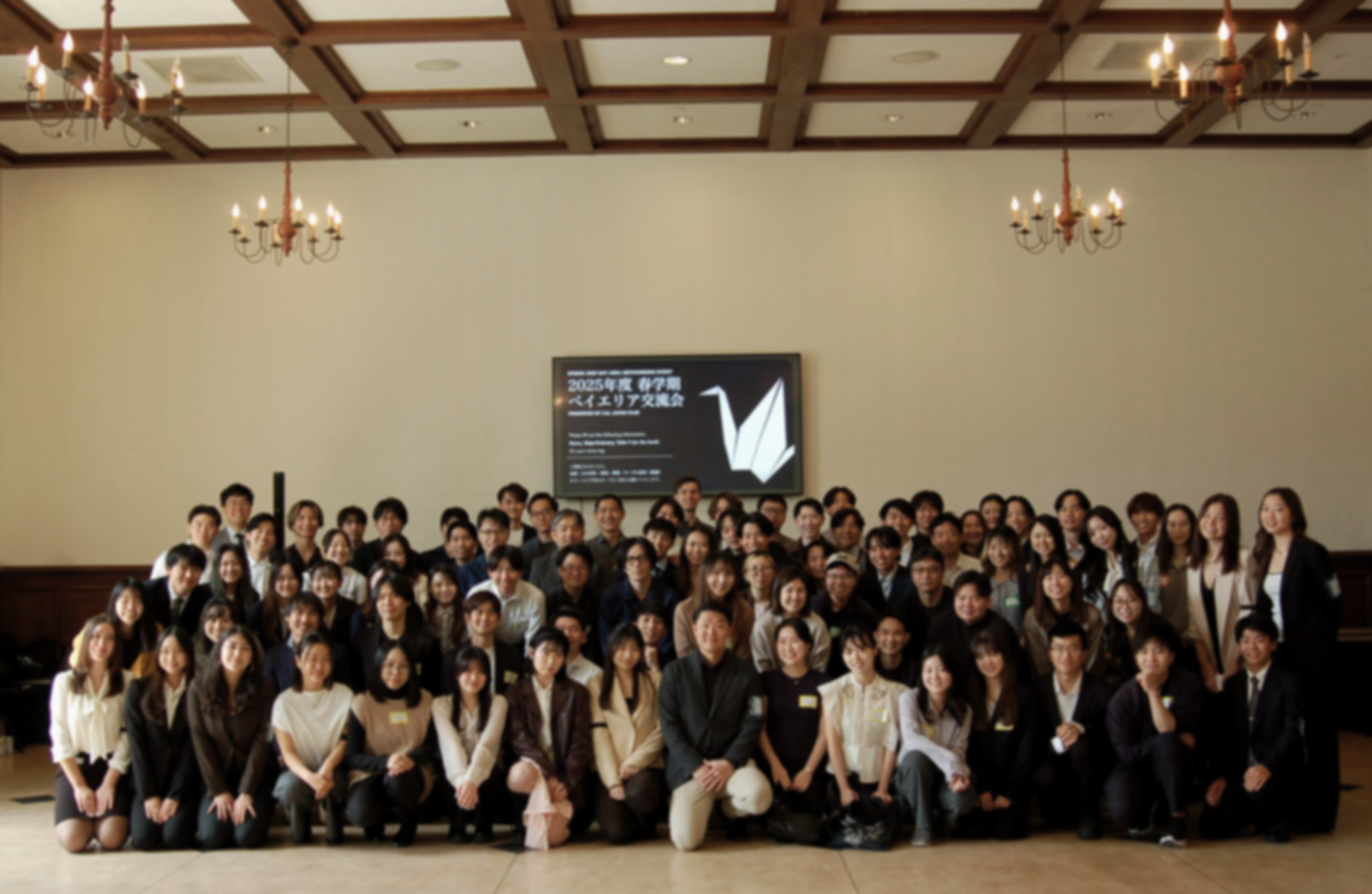 Group photo of people at an indoor event in a large hall with a high ceiling, chandeliers, and a screen displaying Japanese text in the background.