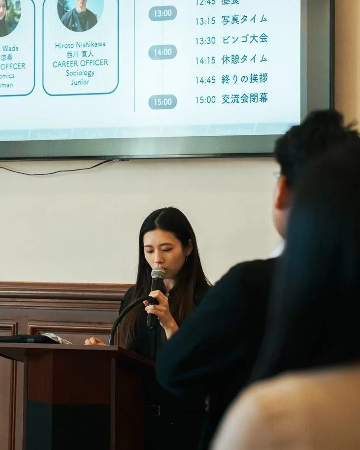 A woman with long dark hair is speaking into a microphone at a small podium in a conference room, with a large schedule or agenda displayed on a projector screen behind her. The schedule includes times and activities written in Japanese.