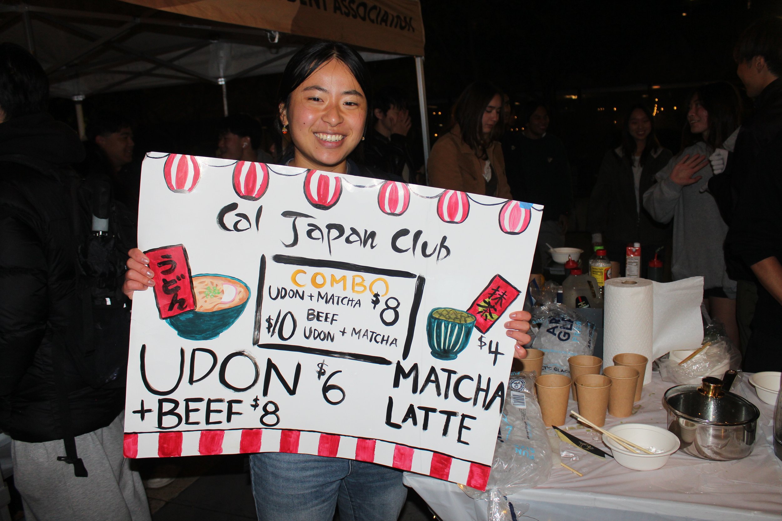 A young woman smiling and holding a large handwritten sign advertising Japanese food items and prices at a night market or outdoor event. The sign features colorful illustrations of ramen bowl and matcha tea.
