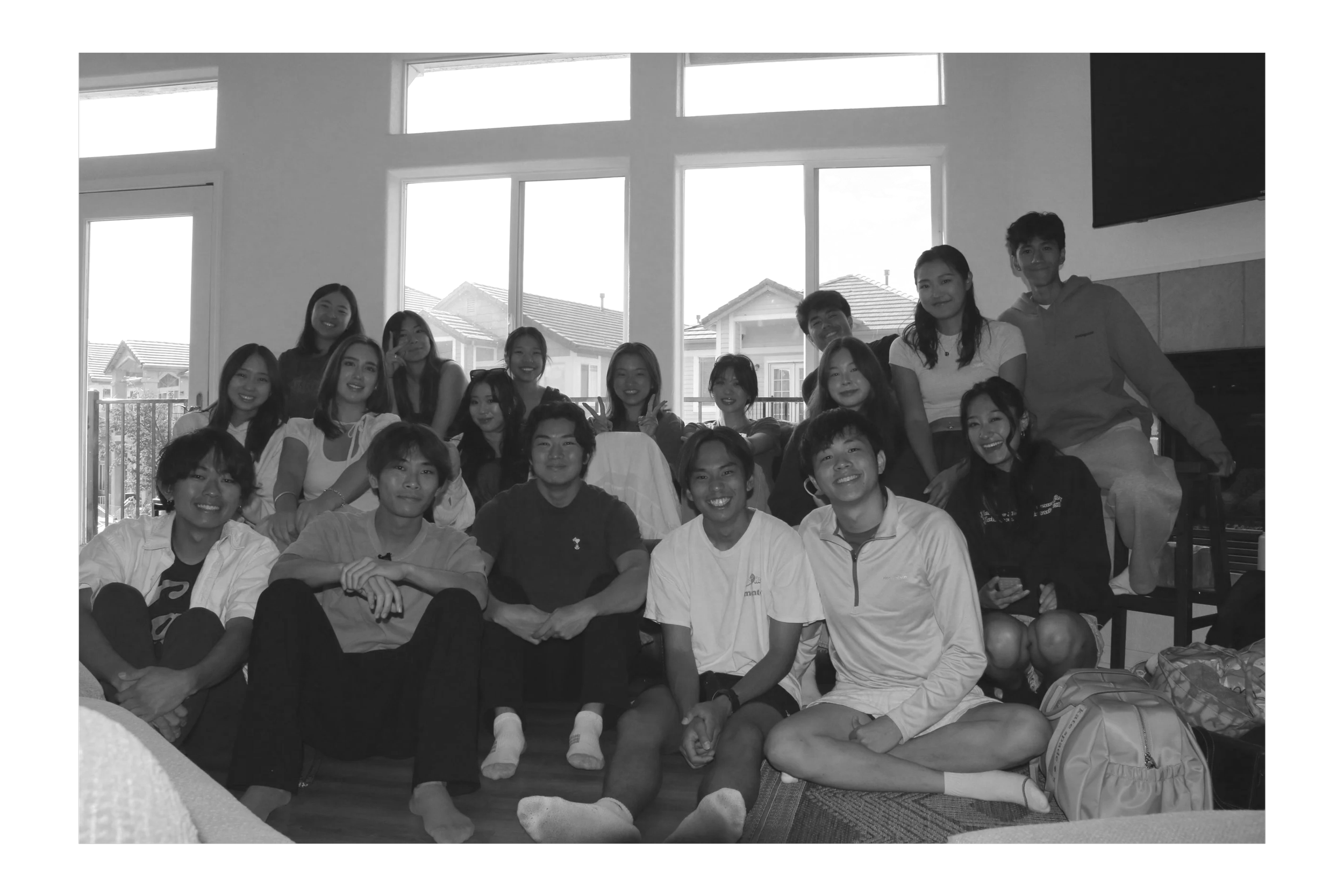 Group of young people smiling indoors, sitting and standing together, with large windows showing houses outside in the background.
