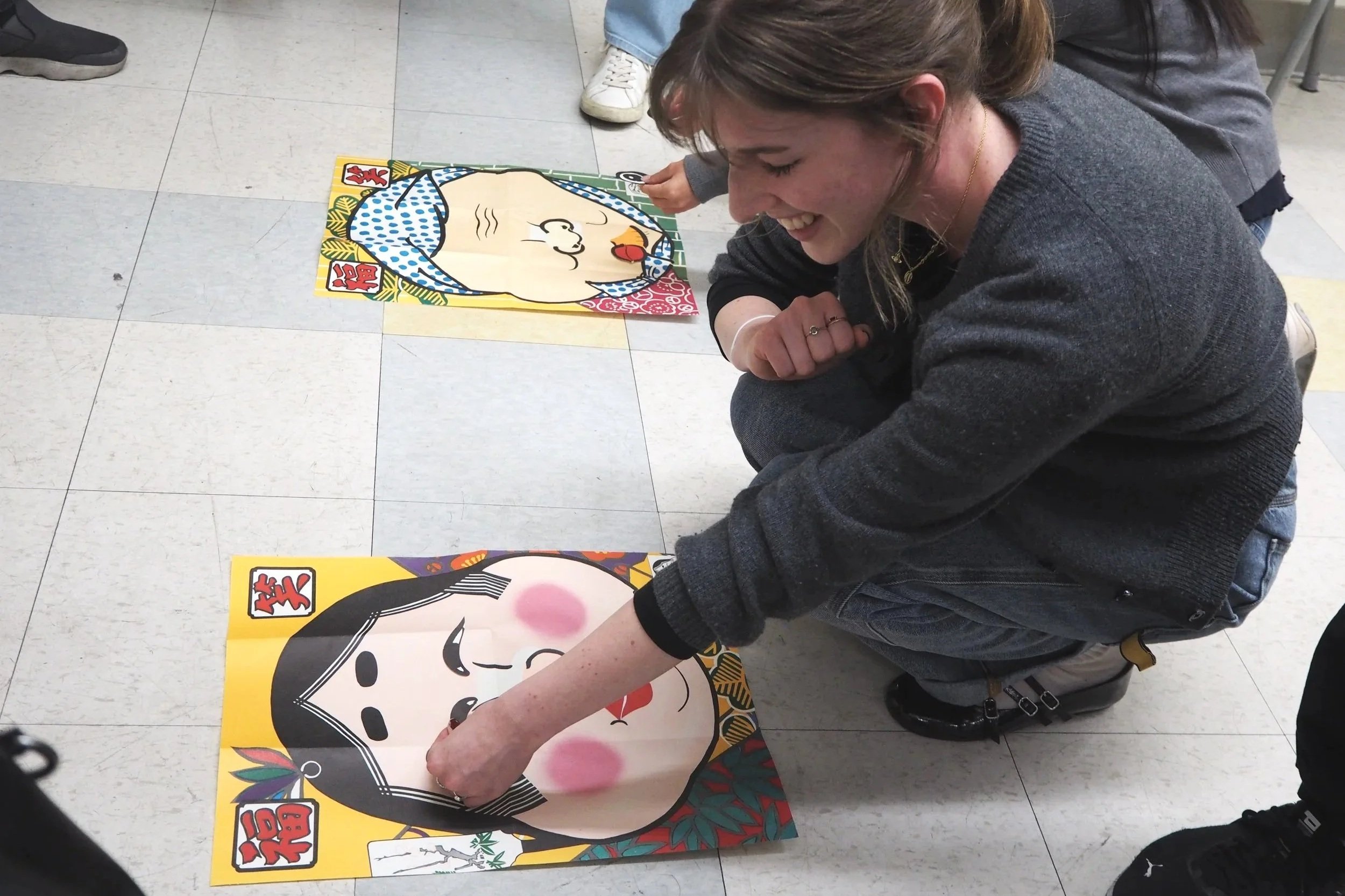 A woman kneeling on the floor of a room playing a mat game, touching the face of a large, cartoon-style playing card with a girl's face on it. There are other similar cards on the floor around her.