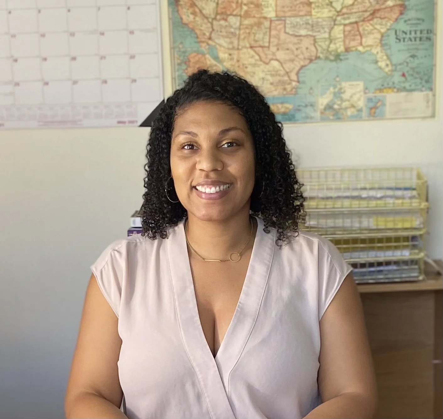 A woman with curly black hair, wearing a beige blouse and jewelry, smiling at the camera in an office with a world map and files in the background.