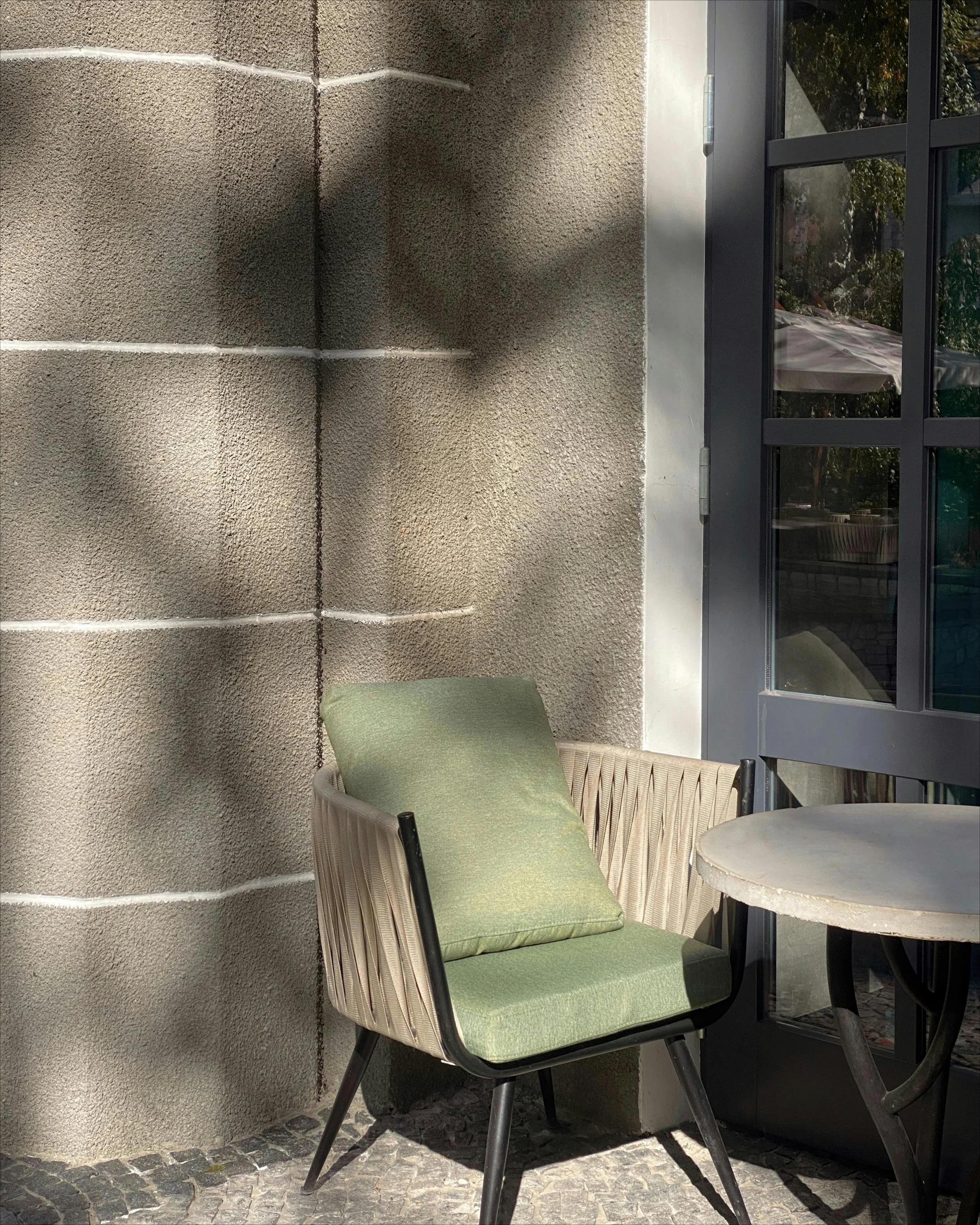 Outdoor patio area with a green cushioned chair against a beige stucco wall, adjacent to a glass door reflecting trees and surroundings.