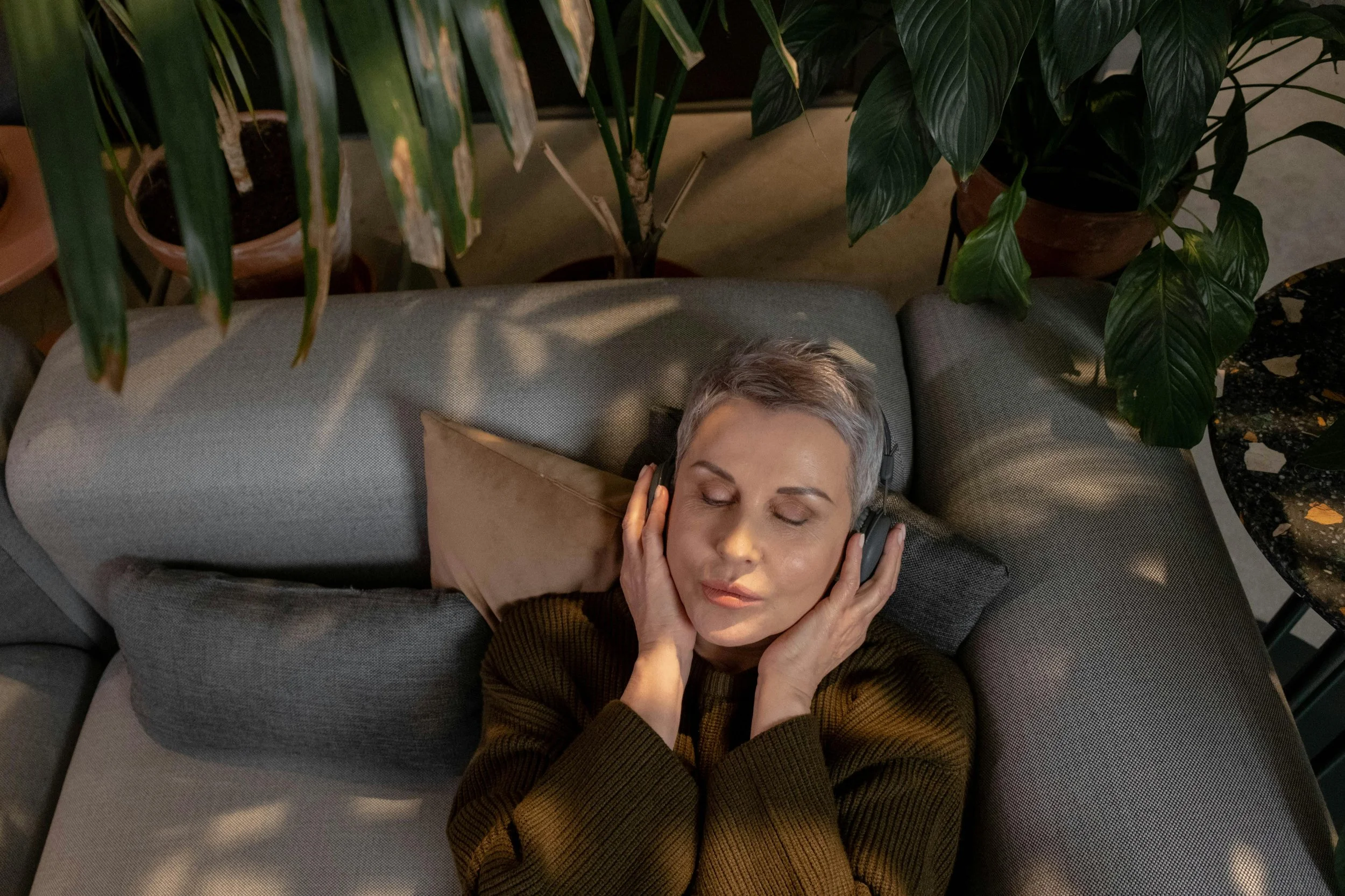 A woman with short gray hair lying on a light gray sofa with her eyes closed, listening to music with over-ear headphones, surrounded by large green plants.