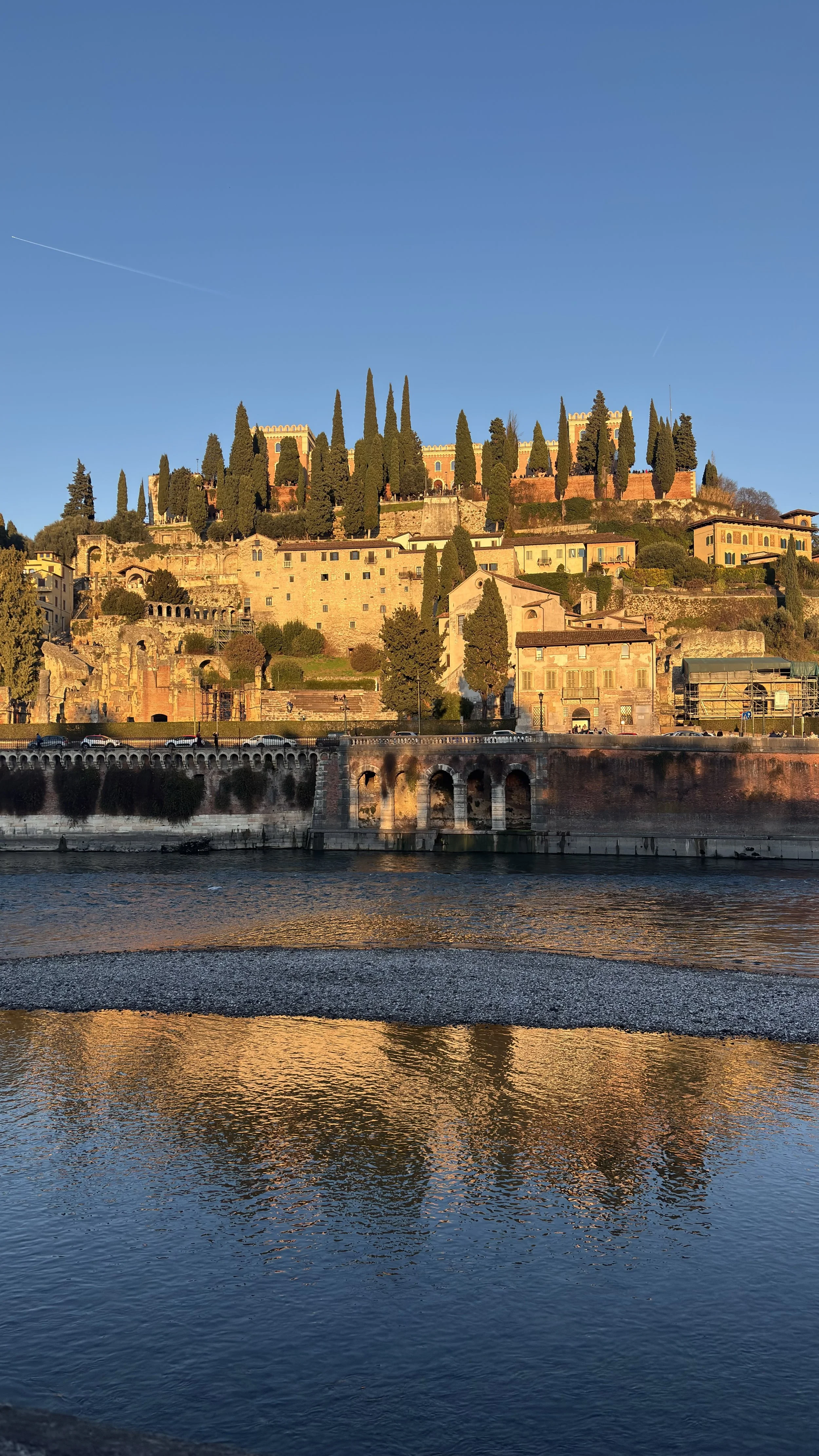 A scenic view of a hillside town with historical buildings and tall cypress trees, reflected in a river at sunset.