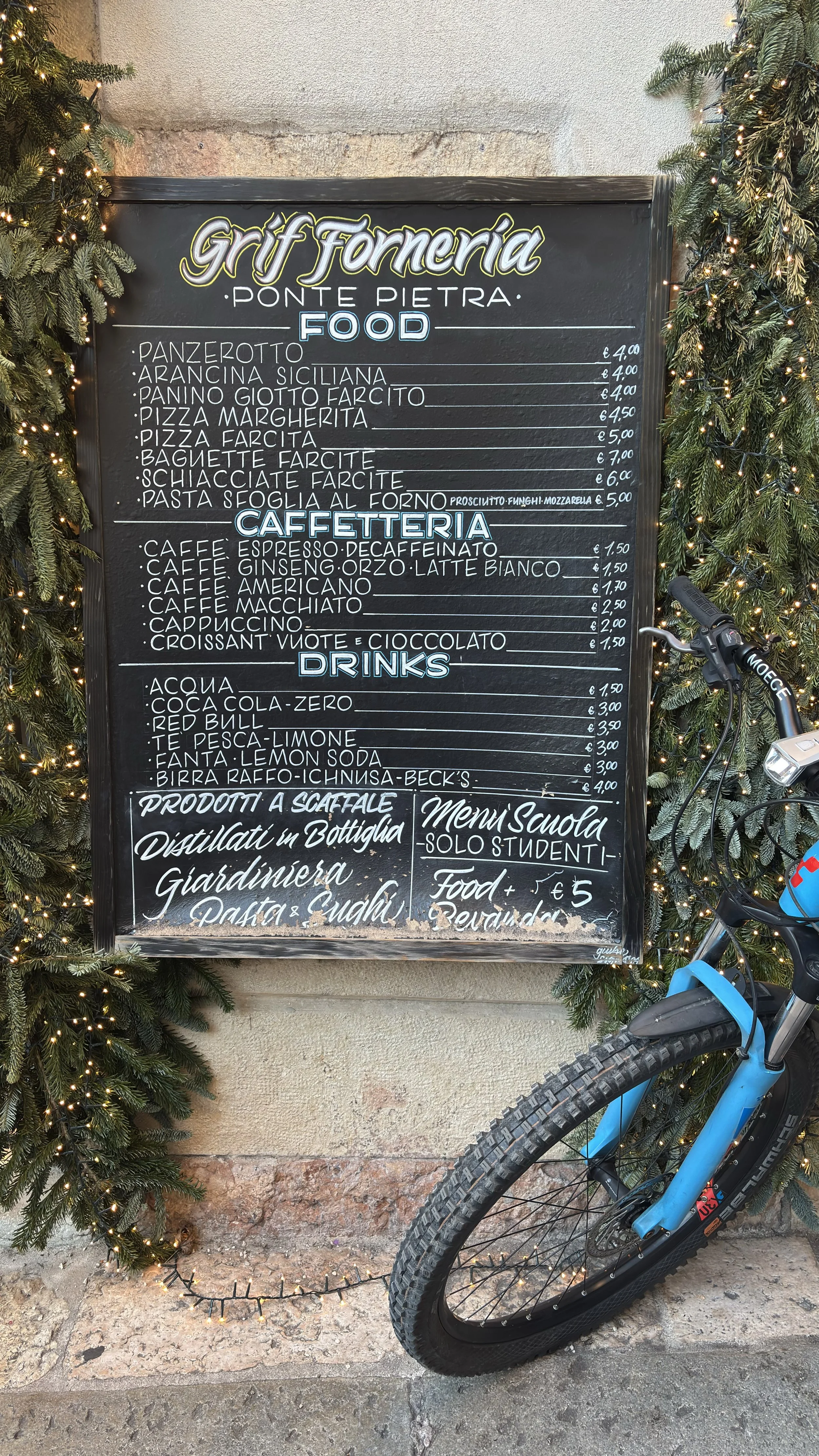 A chalkboard menu outside a restaurant called 'Graft Forneria' on Ponte Pietra, listing food, coffee, drinks, and other items, with some prices in euros. The menu is decorated with evergreen branches and fairy lights, with a bicycle parked nearby.