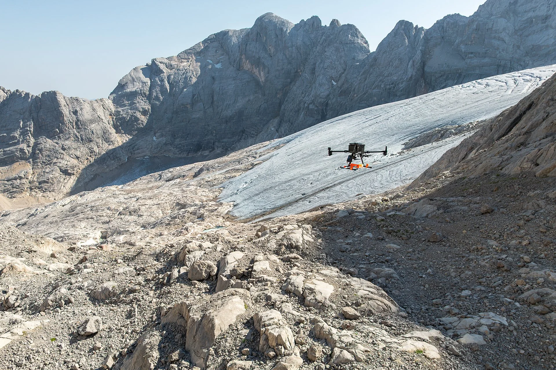 A mountain landscape with rocky terrain, a glacier, and rugged cliffs in the background. A drone is flying over the icy slope.