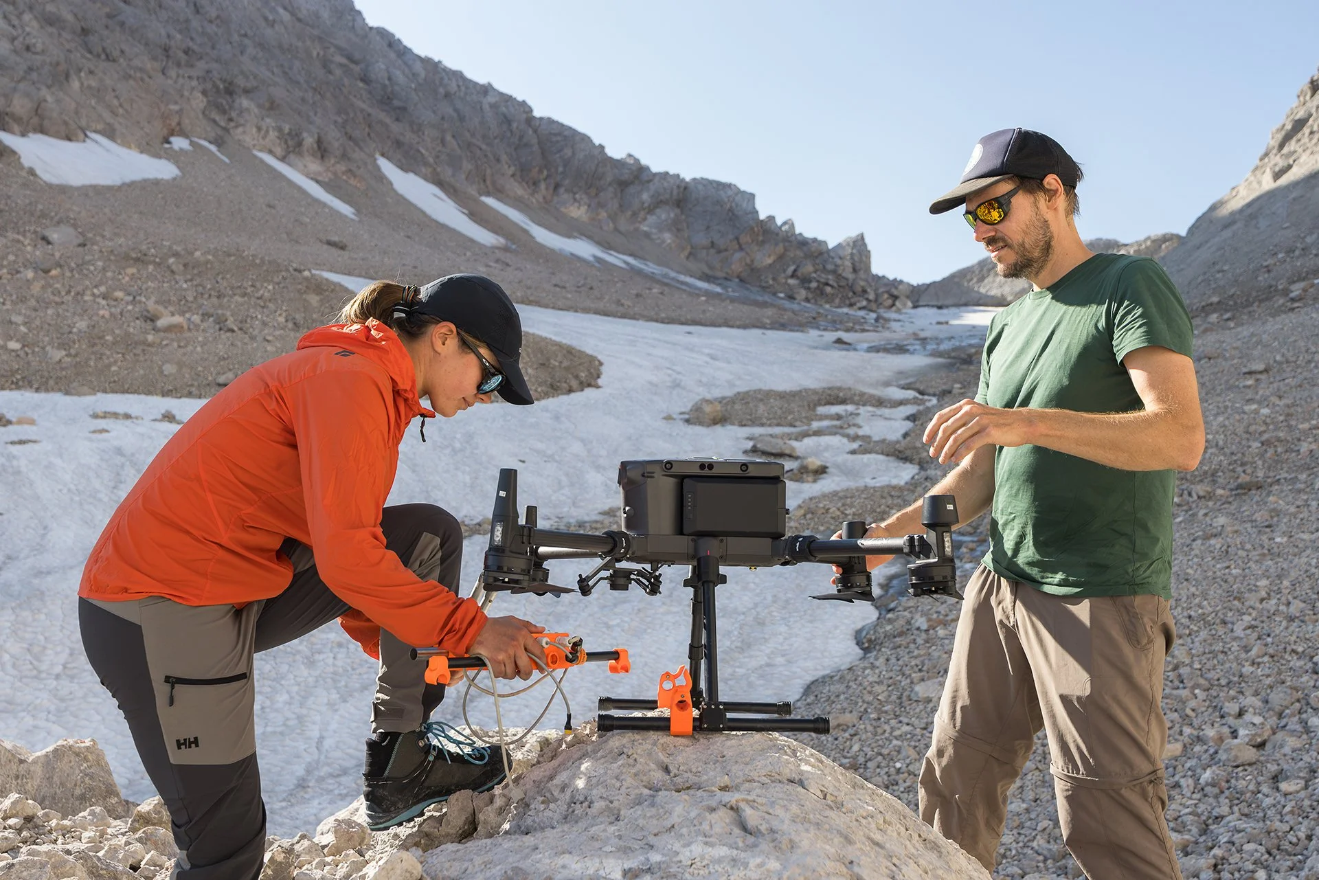 Two scientists set up equipment on rocky, snow-covered terrain in mountainous area with clear sky.