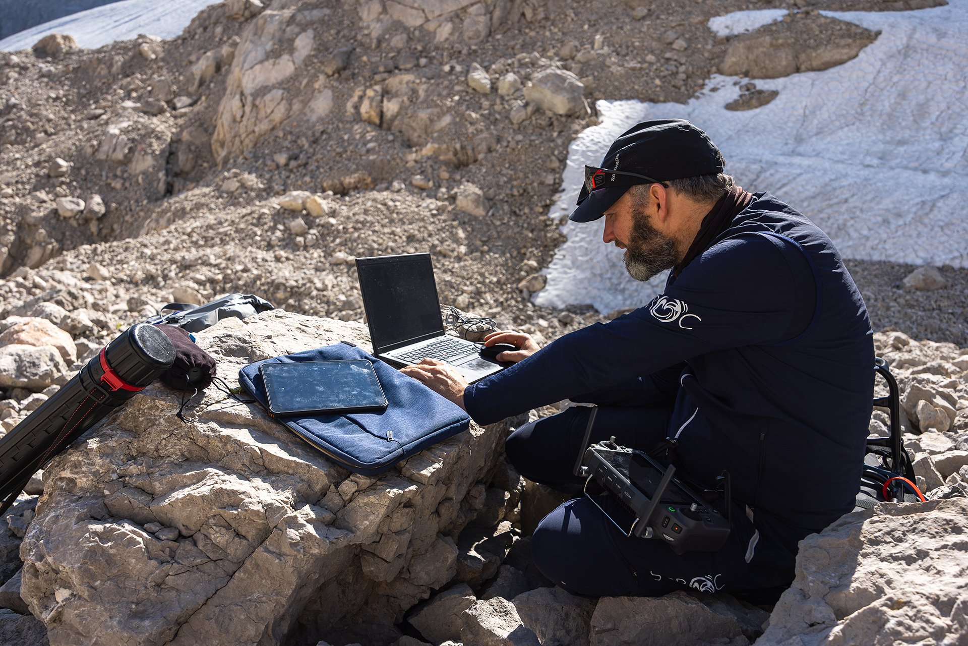 A man with a beard, wearing a black cap and dark outdoor clothing, is sitting on rocks in a mountainous area with snow patches. He is working on a laptop with various electronic devices around, including a tablet, a black case, and a camera with a tr