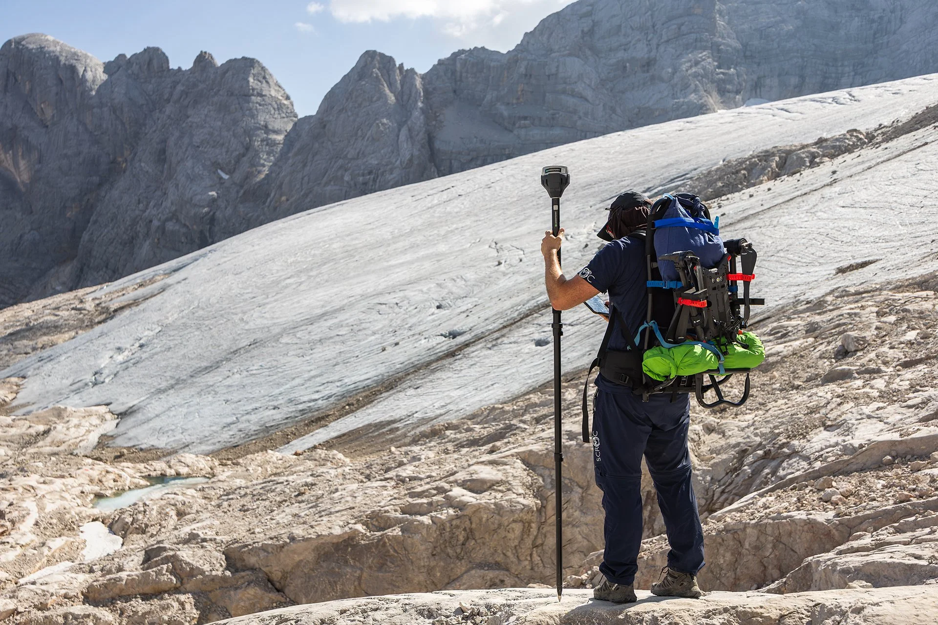A male hiker standing on a snow-covered mountain trail with rocky terrain, carrying a large backpack with outdoor gear, holding a walking stick, and looking at a map against a backdrop of rugged mountain peaks.