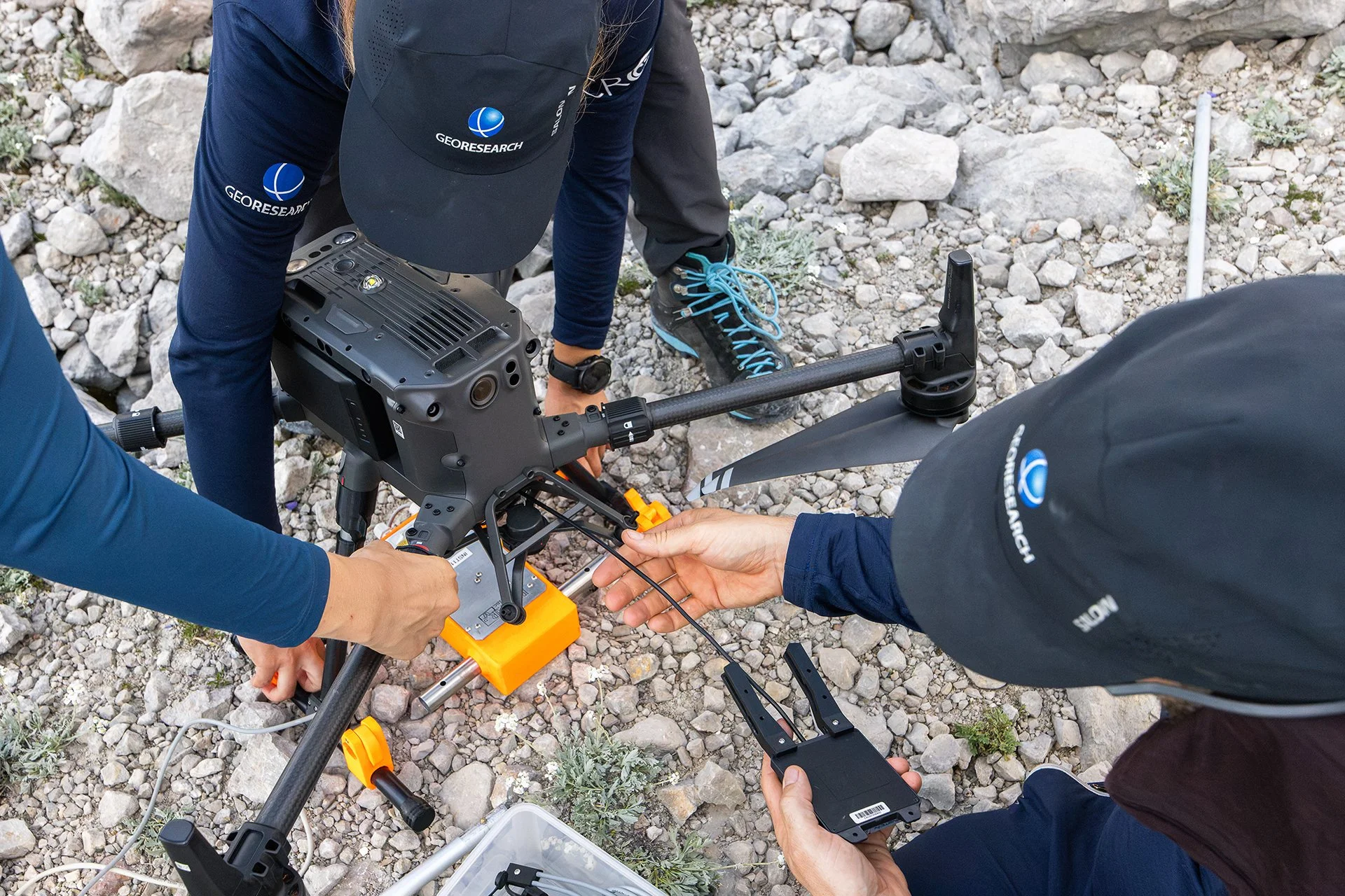 Scientists or researchers working with a drone or unmanned aerial vehicle on rocky terrain, possibly in a desert or mountainous area, with equipment and wires around.