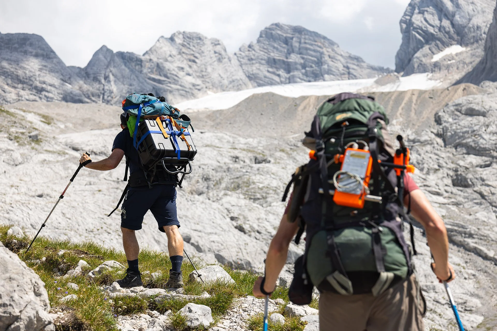 Two hikers with backpacks and trekking poles walking in a mountainous terrain with rocky slopes and snow-capped peaks in the background.