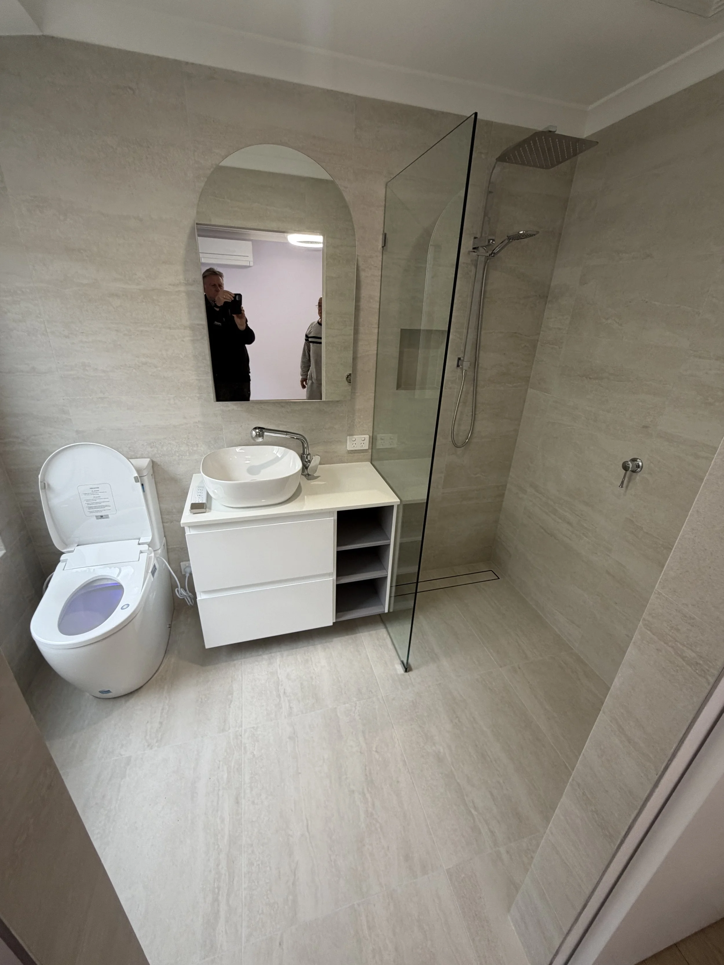 A modern bathroom with a toilet, sink with a counter, mirror, and a walk-in shower enclosed by a glass partition. The bathroom has light-colored tiled walls and flooring.