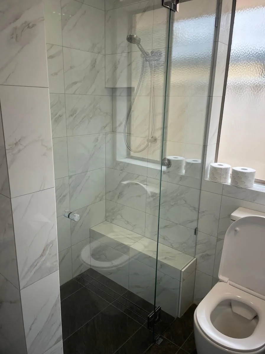 Shower area with marble tiles, a glass partition, a handheld showerhead, a built-in bench, and toilet with toilet paper rolls on the window sill.