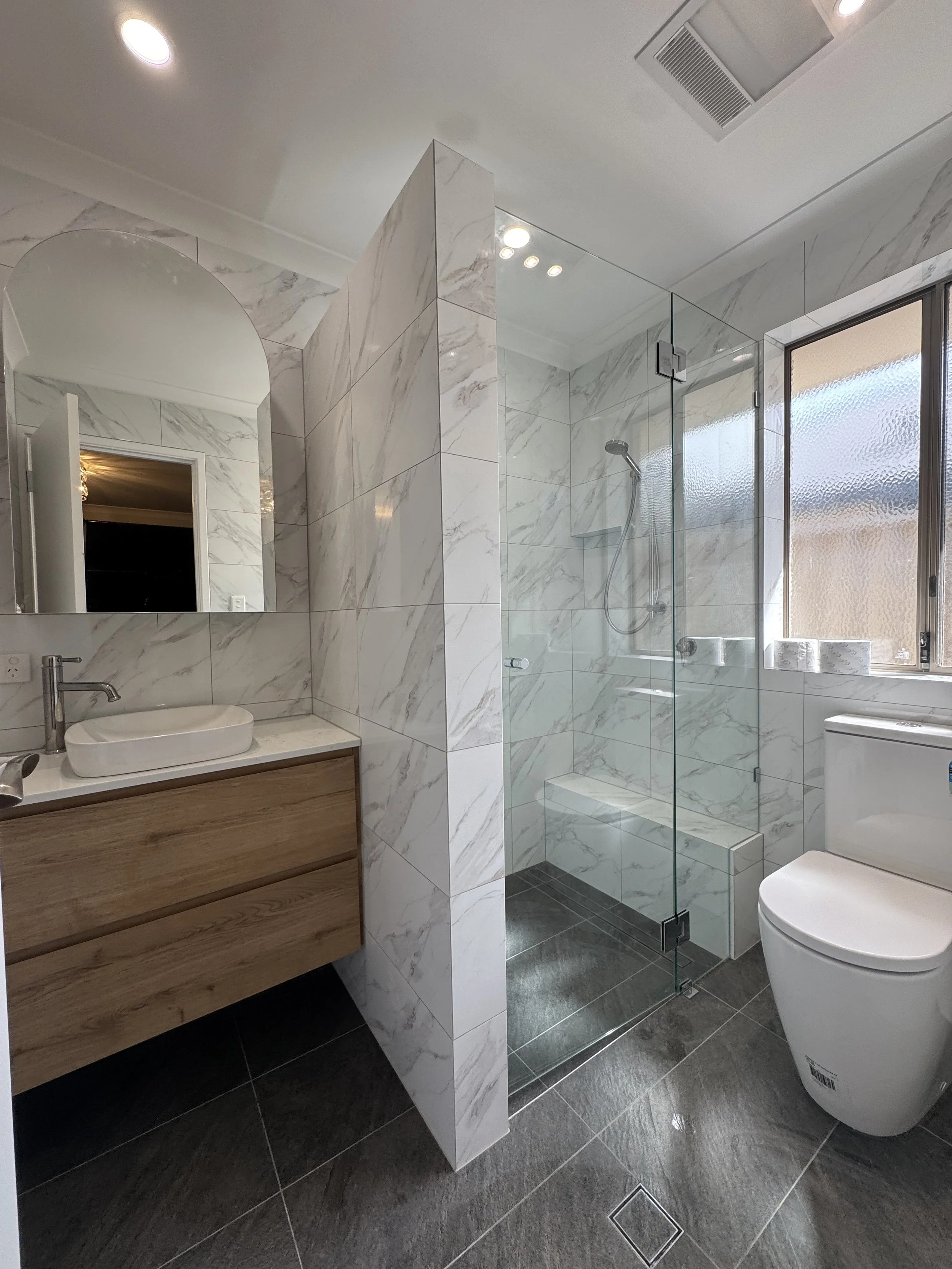 Modern bathroom with a white marble shower enclosure, a wooden vanity with a vessel sink, a large mirror, and a white toilet. The bathroom has a frosted window and dark tiled flooring.