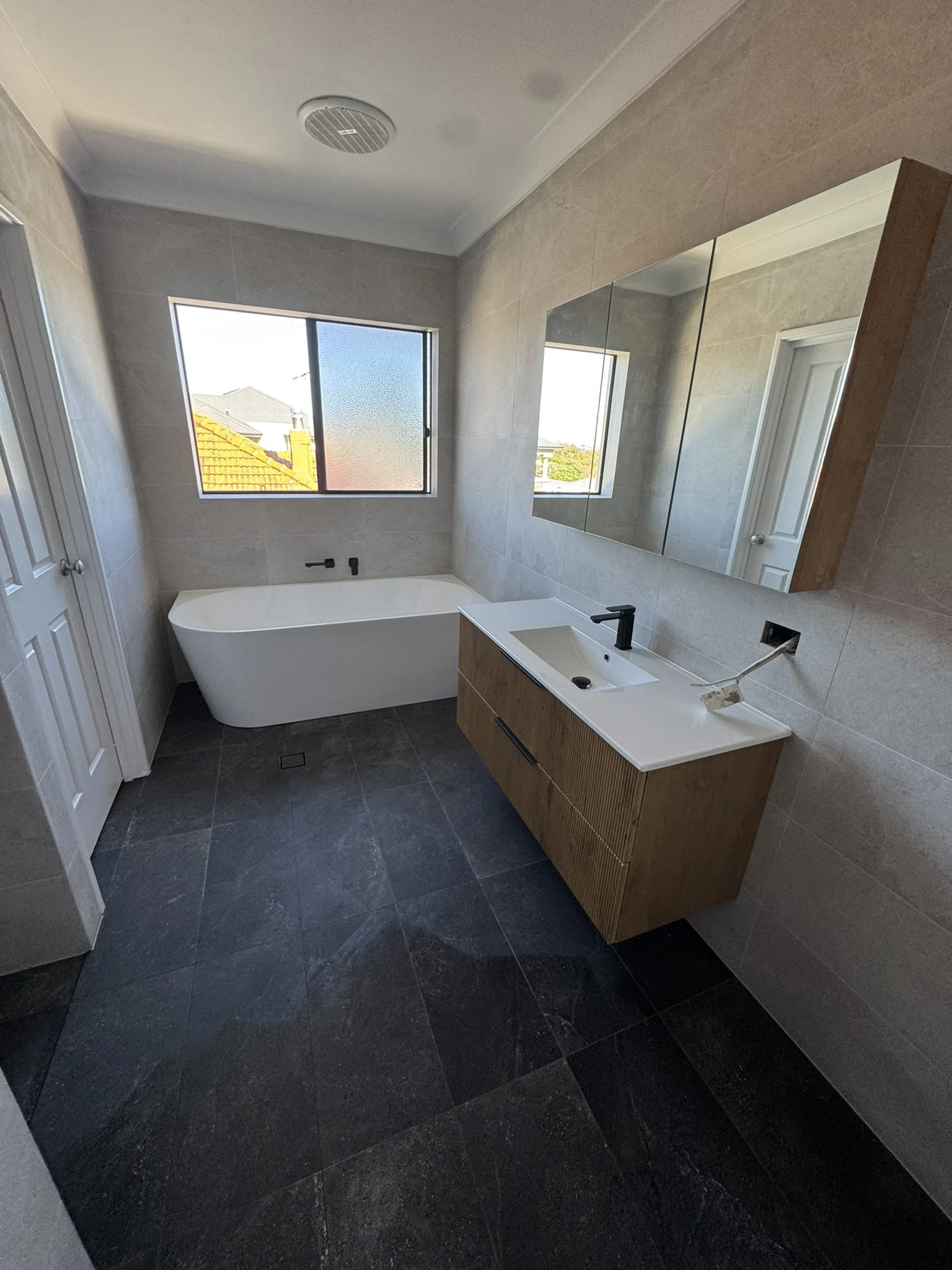Modern bathroom with a rectangular bathtub near a window, a wooden floating vanity with an integrated sink, and a large mirror, all with beige tiled walls and dark tiled flooring.