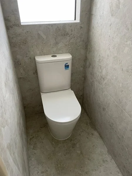 A white toilet in a small bathroom with gray stone walls and floor, and a window above the toilet.