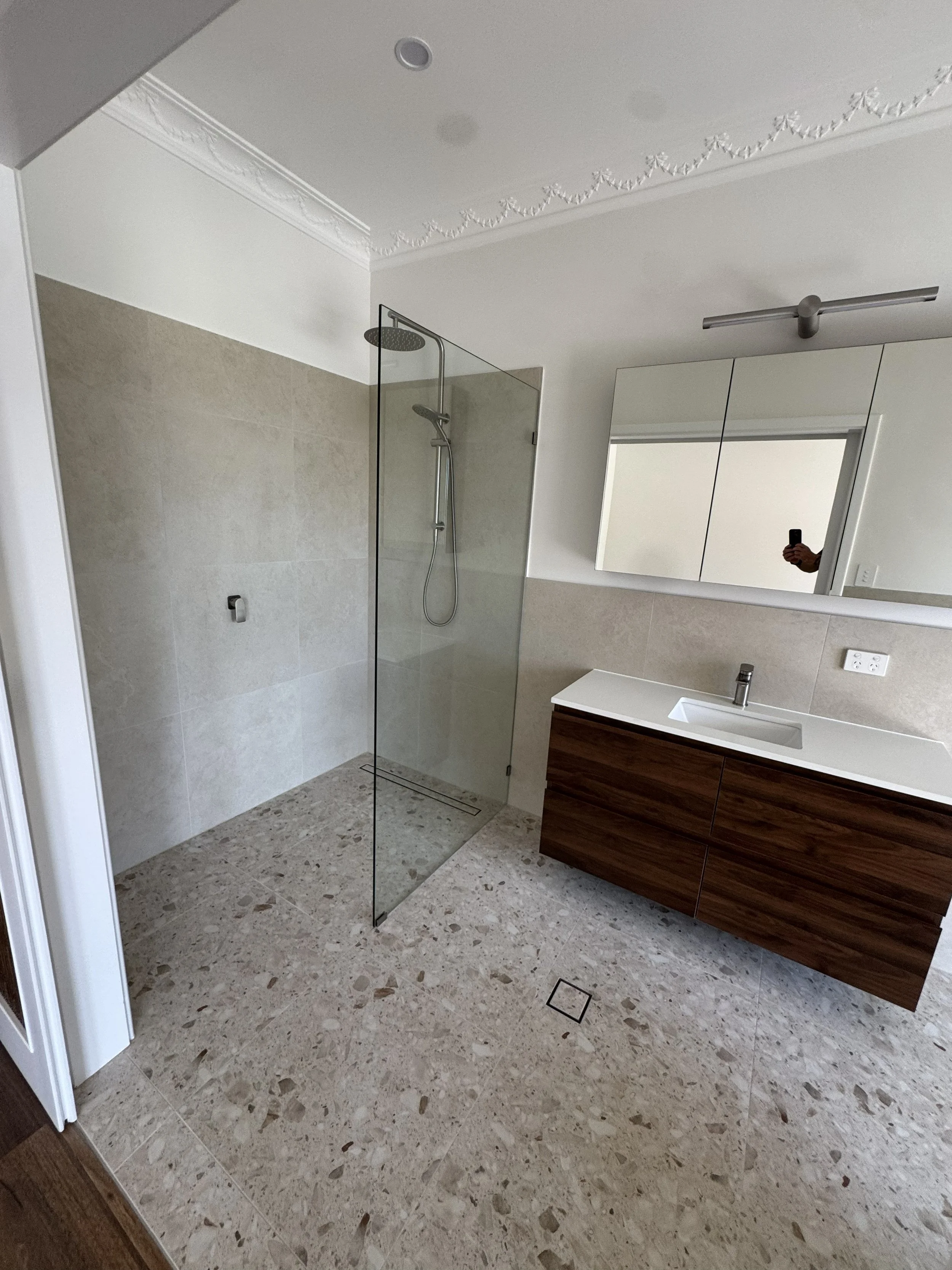 A modern bathroom with beige tiled floor and walls, a glass shower enclosure, a white sink with a wooden cabinet, a mirrored cabinet, and a ceiling fan.