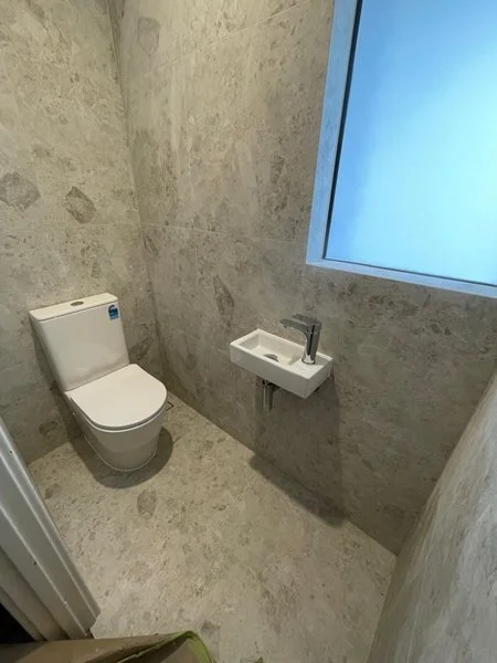 Small bathroom with a toilet, a tiny sink, and a frosted window on a beige marble wall.