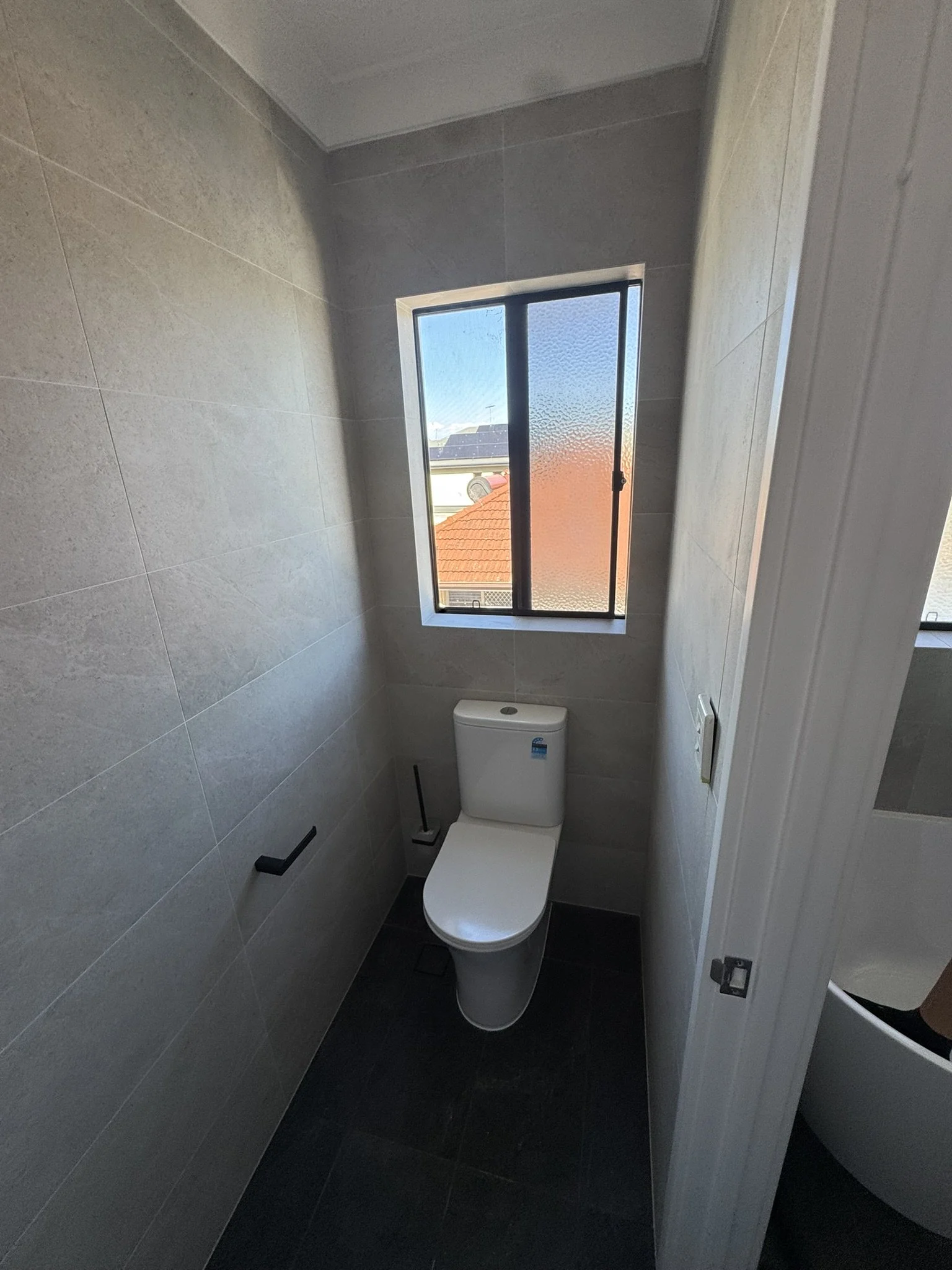Small bathroom with a white toilet, a window with frosted and clear glass, beige tiled walls, and dark tiled floor.