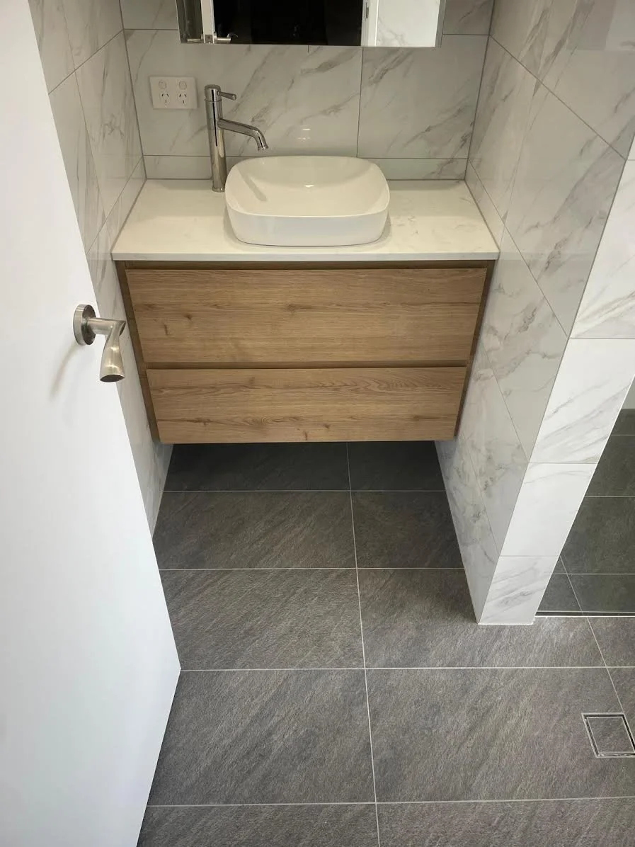 A modern bathroom vanity with a round vessel sink, a tall faucet, and a wooden cabinet, set against tiled marble walls and dark floor tiles.
