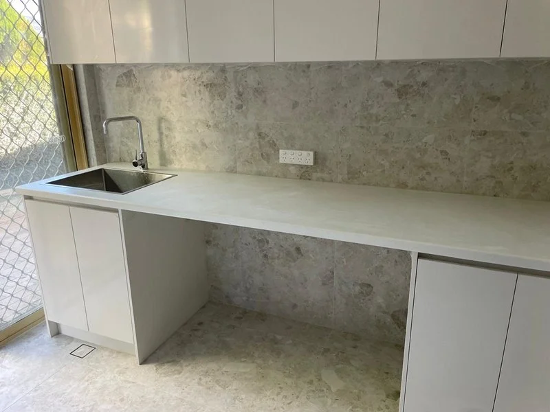 Minimalist kitchen with a white countertop, a sink with a chrome faucet, white cabinets above and below, a textured beige backsplash, a power outlet, and a sliding glass door with a screen.