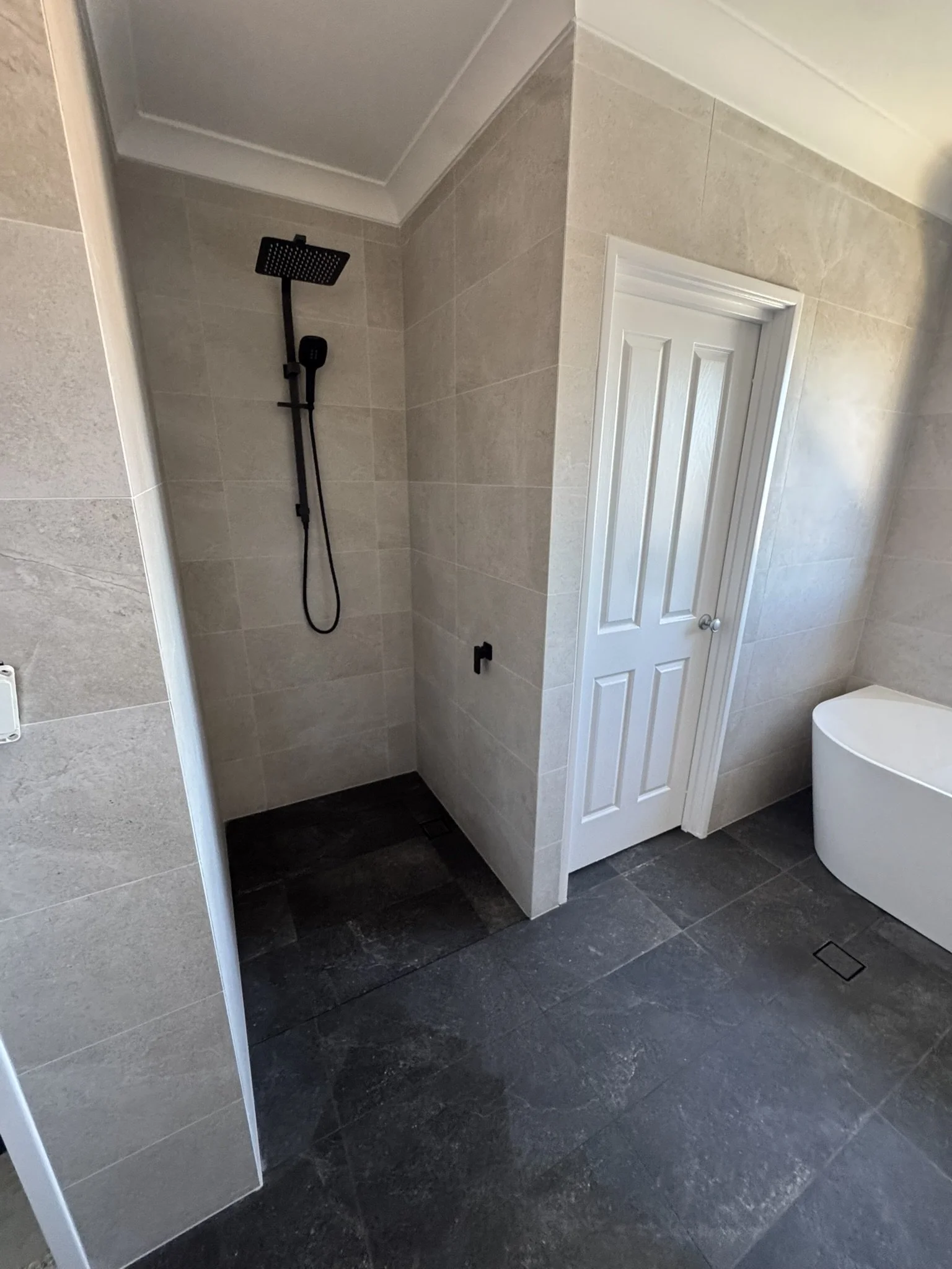 Bathroom with a walk-in shower with black fixtures and a white door, beige tiled walls, dark tiled floor, and a white bathtub.
