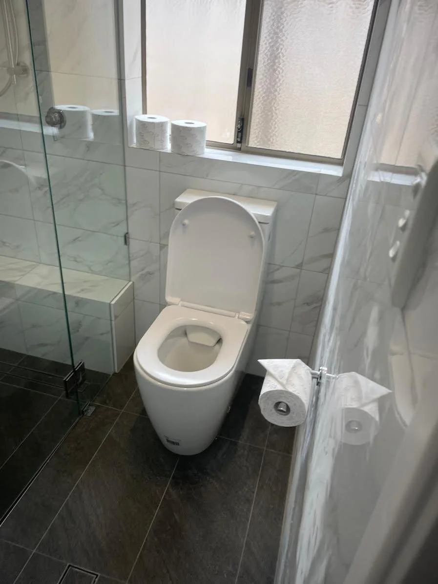 Bathroom with a white toilet, three rolls of toilet paper on the window sill, a glass shower door, and black and white tiled walls.
