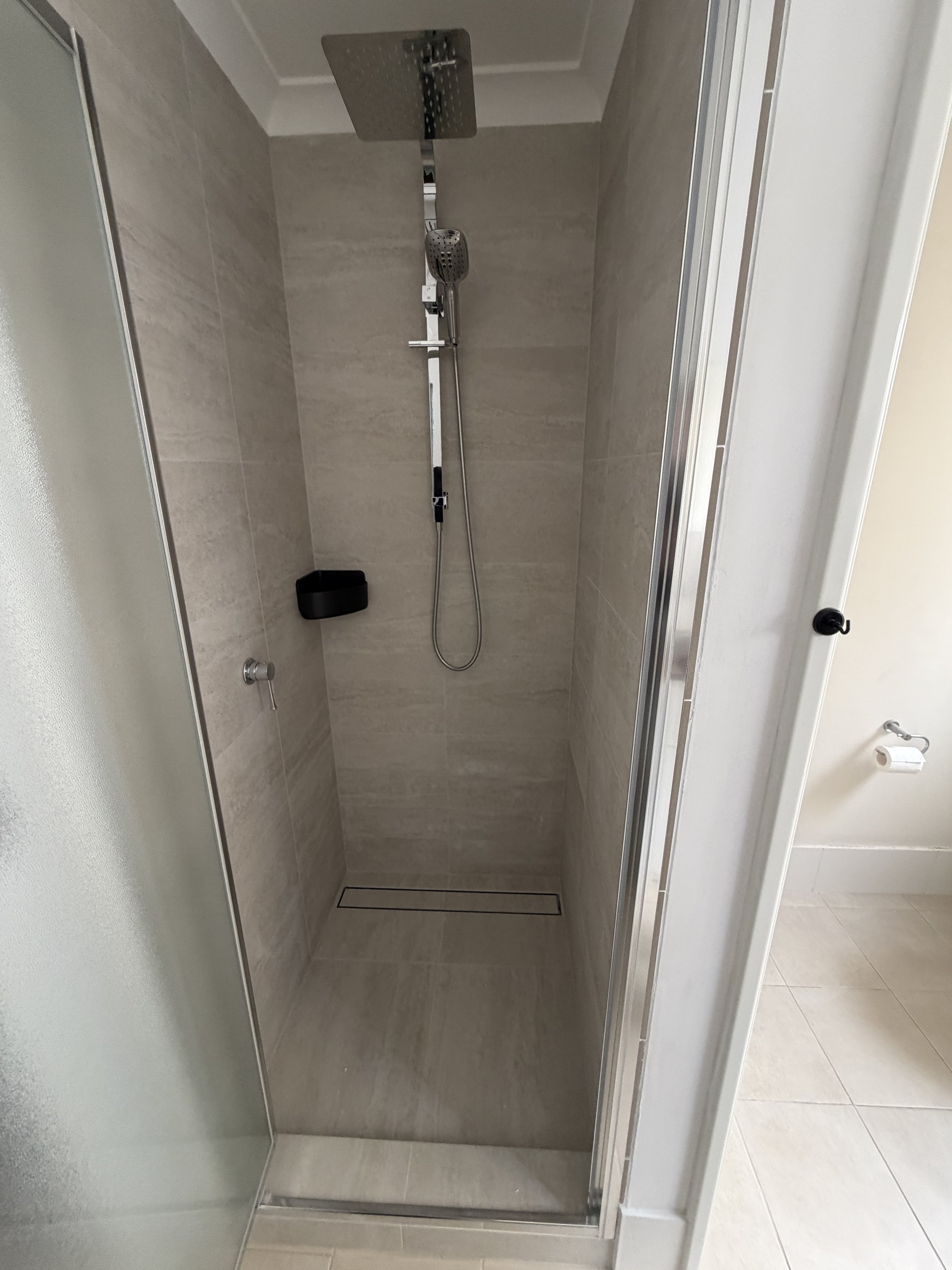 A shower with a square rainfall showerhead and handheld showerhead, beige tiled walls, a black soap dish, and a linear drain on the floor.