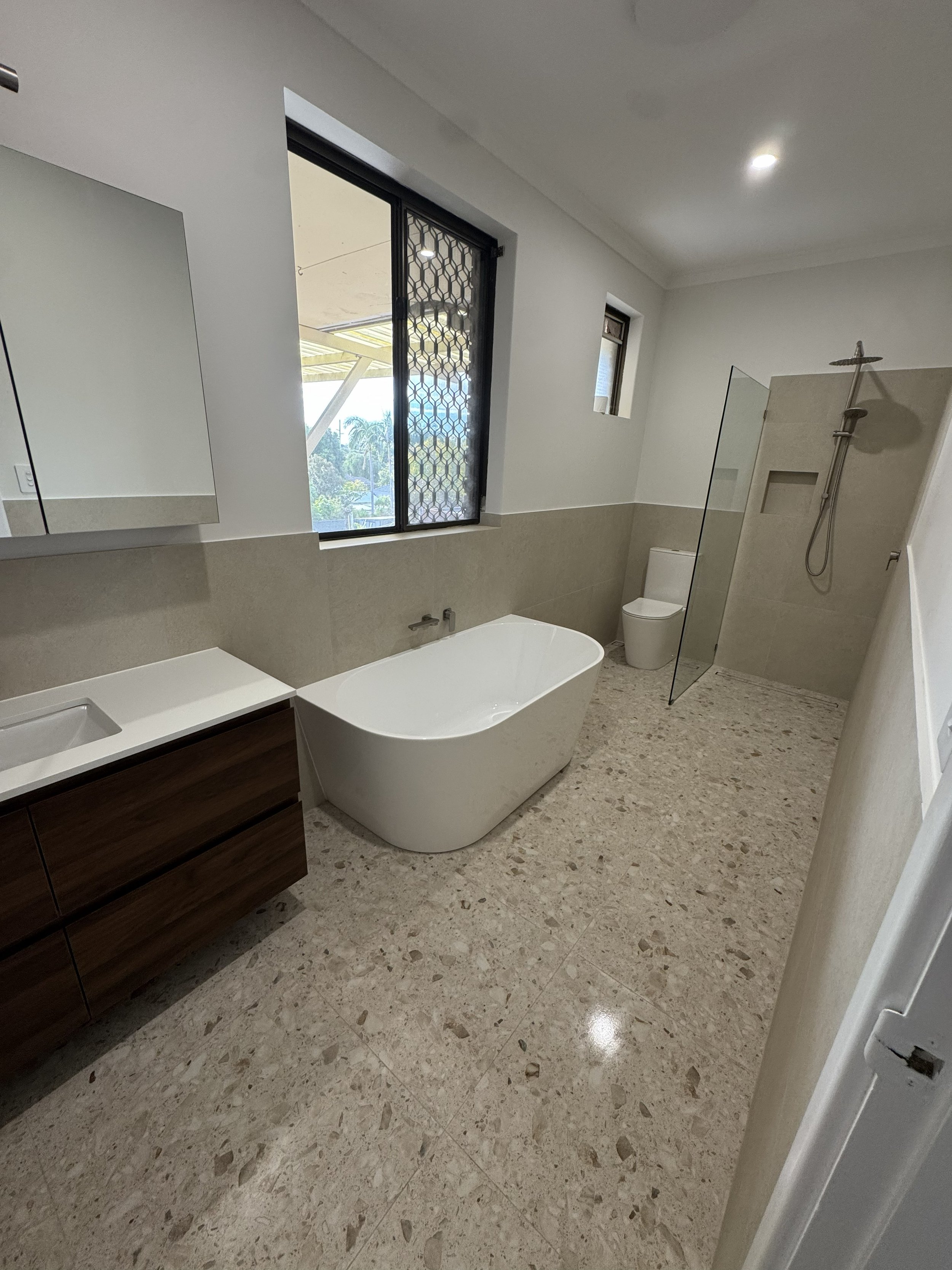 A modern bathroom with a freestanding bathtub, a vanity with a sink, a wall-mounted mirror, a toilet, a walk-in shower with glass partition, and windows providing natural light.