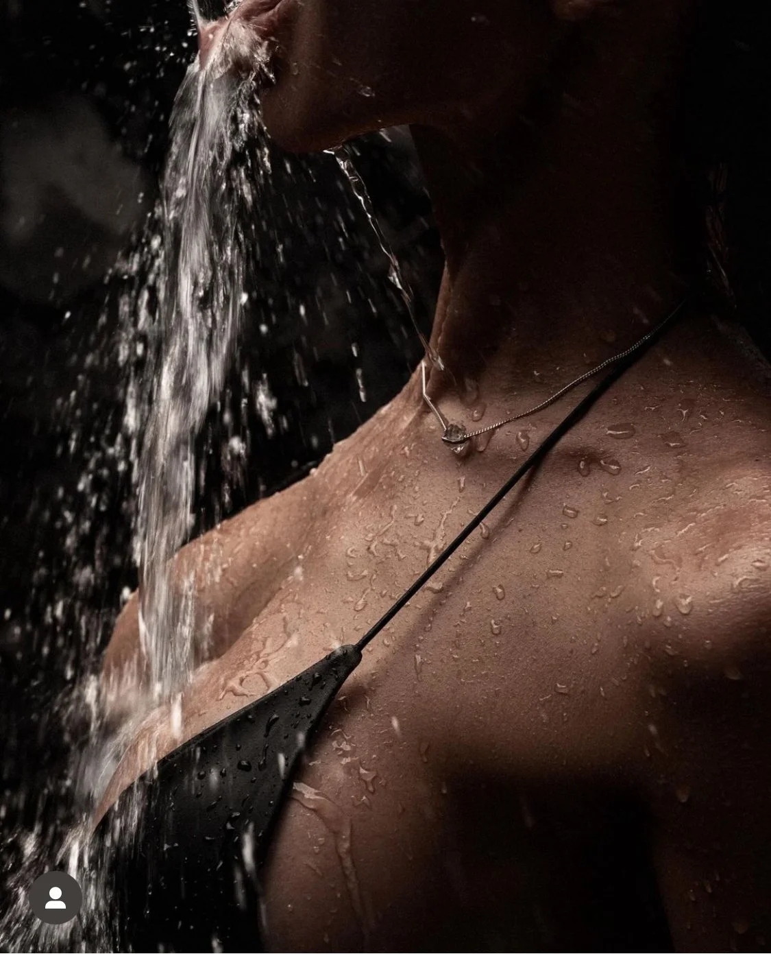 A woman in a dark setting wearing a black swimsuit with water flowing over her body, focusing on her neck and shoulders.