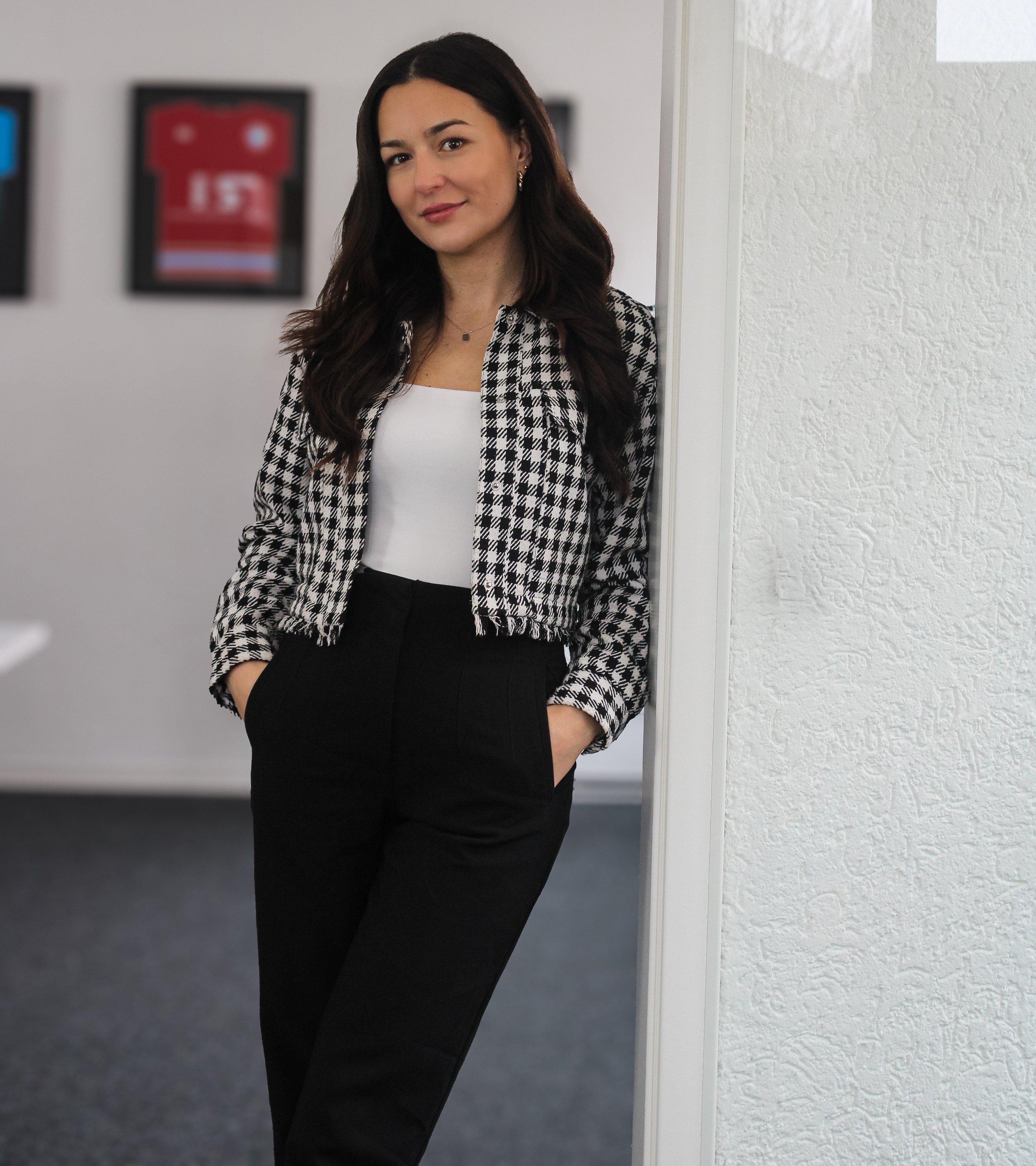 Junge Frau mit dunklen Haaren, in Schwarz-Weiß-Checkered-Jacke und schwarzer Hose, lehnt an einer Wand in einem Büro.
