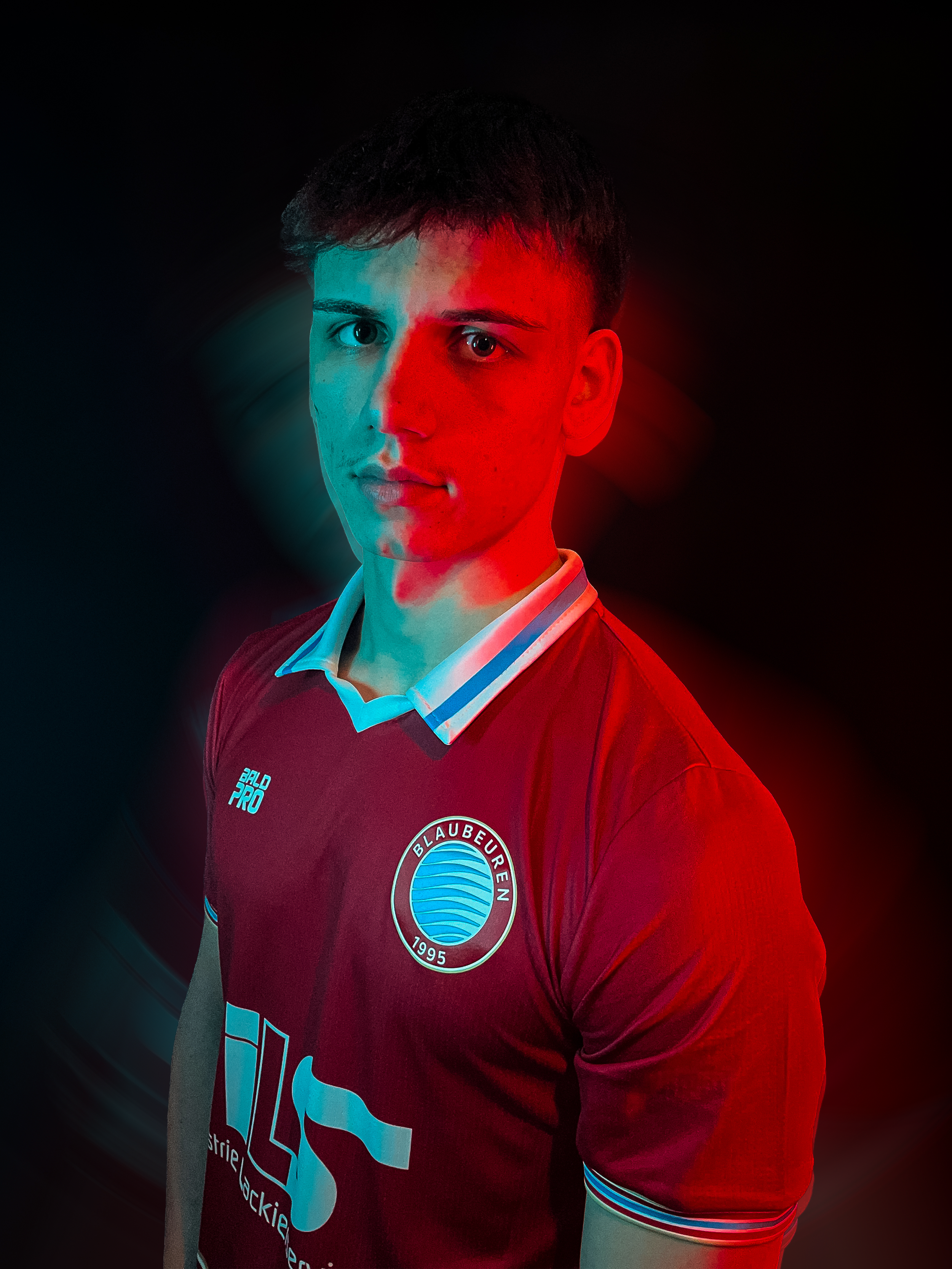 Junger Mann trägt ein Rot-weißes Fußballtrikot mit Blaubeuren-Logo, dunklem Hintergrund, farbigem Licht.