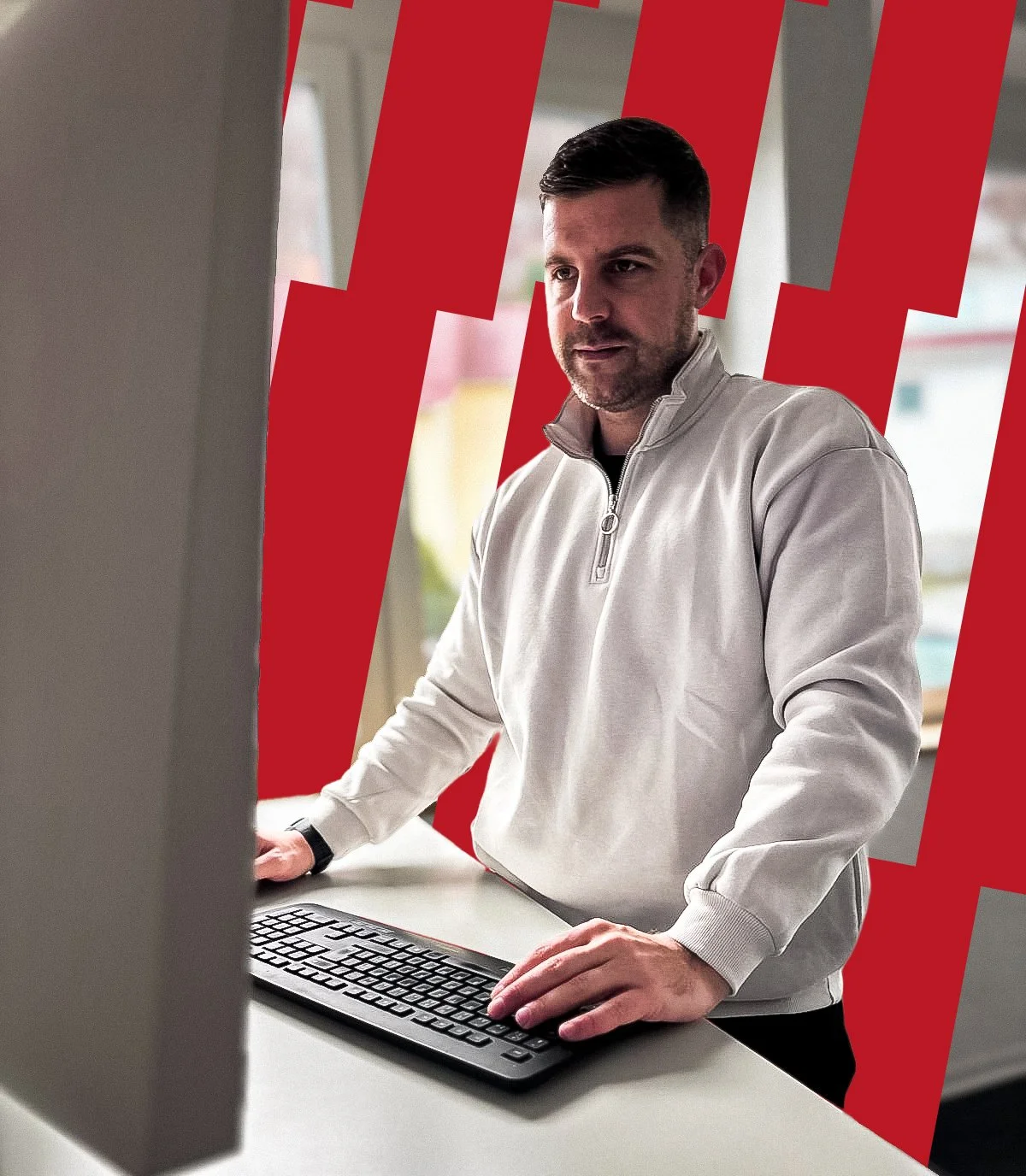 Mann arbeitet an einem Computer in einem Raum mit großen Fenstern und rotem Hintergrundmuster.