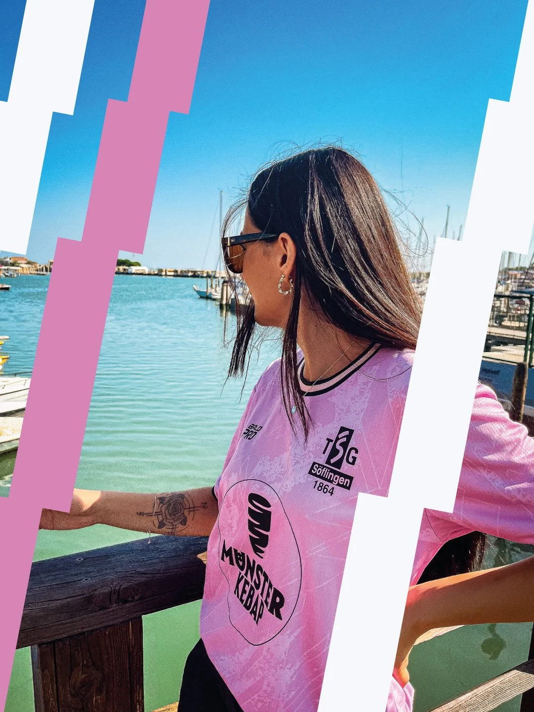 Frau mit dunklen Haaren, Sonnenbrille und Tattoo auf dem Arm steht an einem Hafen mit Booten, trägt ein pinkes T-Shirt mit schwarzen Schriftzügen und steht an einem Holzzaun.