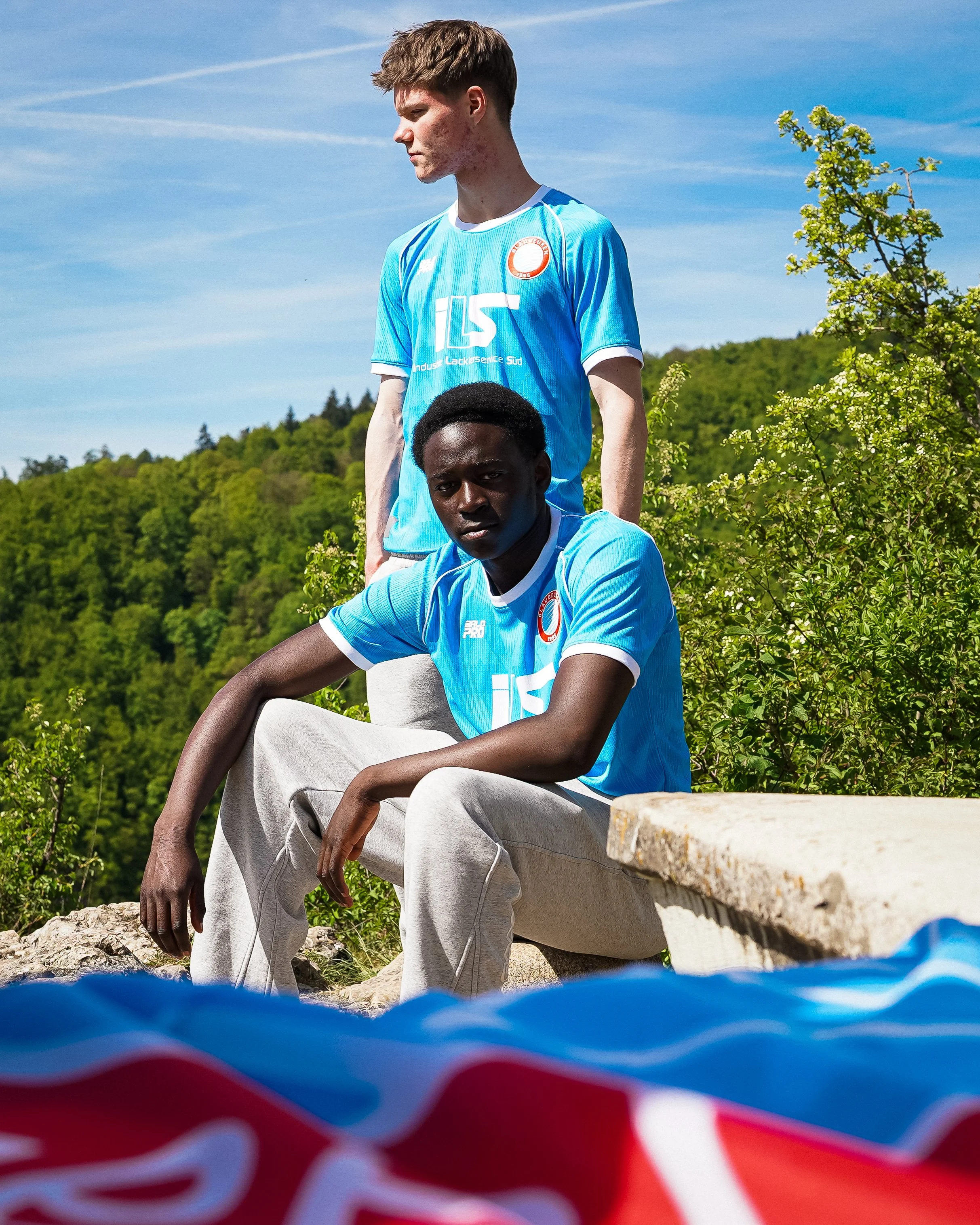 Zwei junge Männer in blauen Trikots, einer sitzend, einer stehend, im Freien in der Natur bei sonnigem Wetter, mit Bäumen und Himmel im Hintergrund.