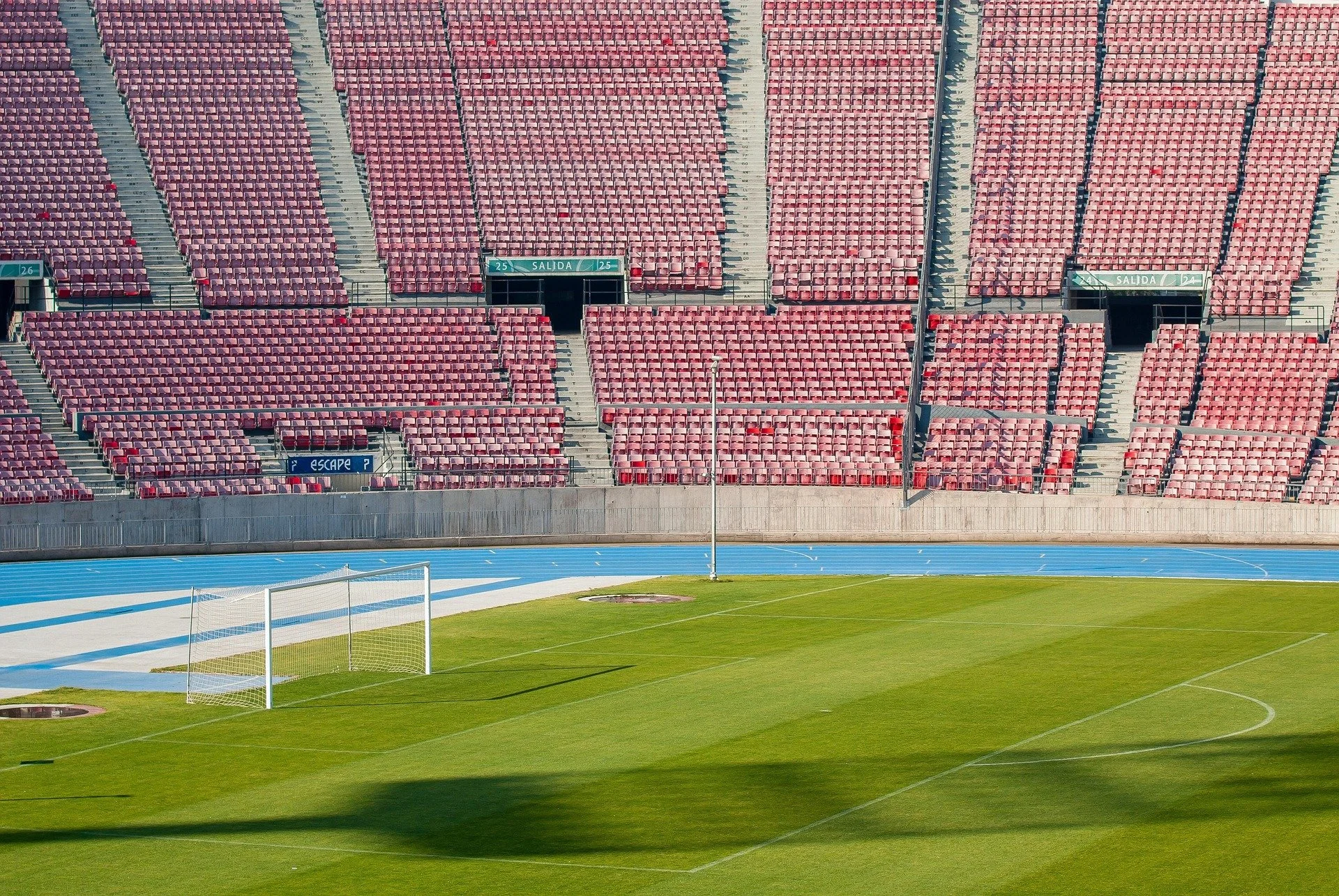 Leeres Fußballstadion mit grüner Spielfläche, Tor und laufbahn sowie roten Sitzplätzen in den Tribünen.