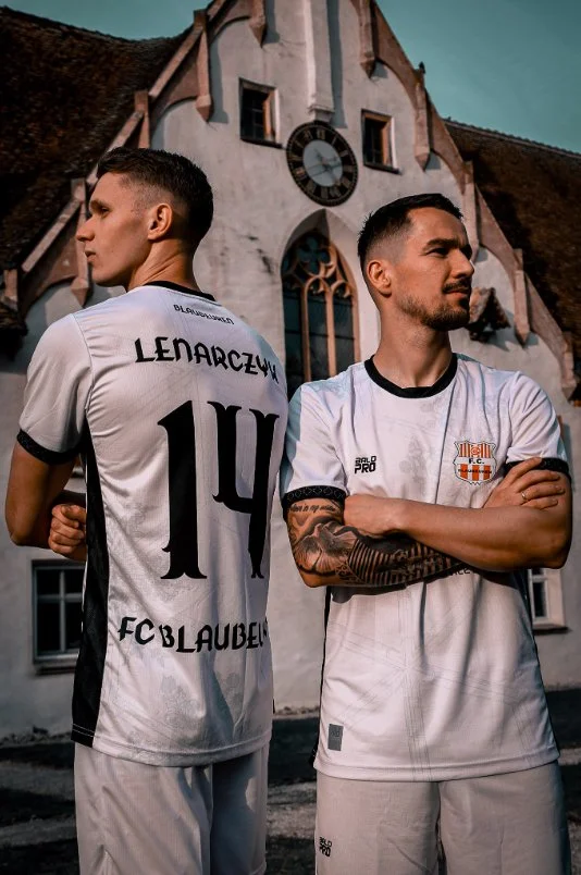 Zwei junge Männer in Fußballtrikots vor einer alten Kirche mit einem Zifferblatt-Uhrturm, während die Sonne scheint.