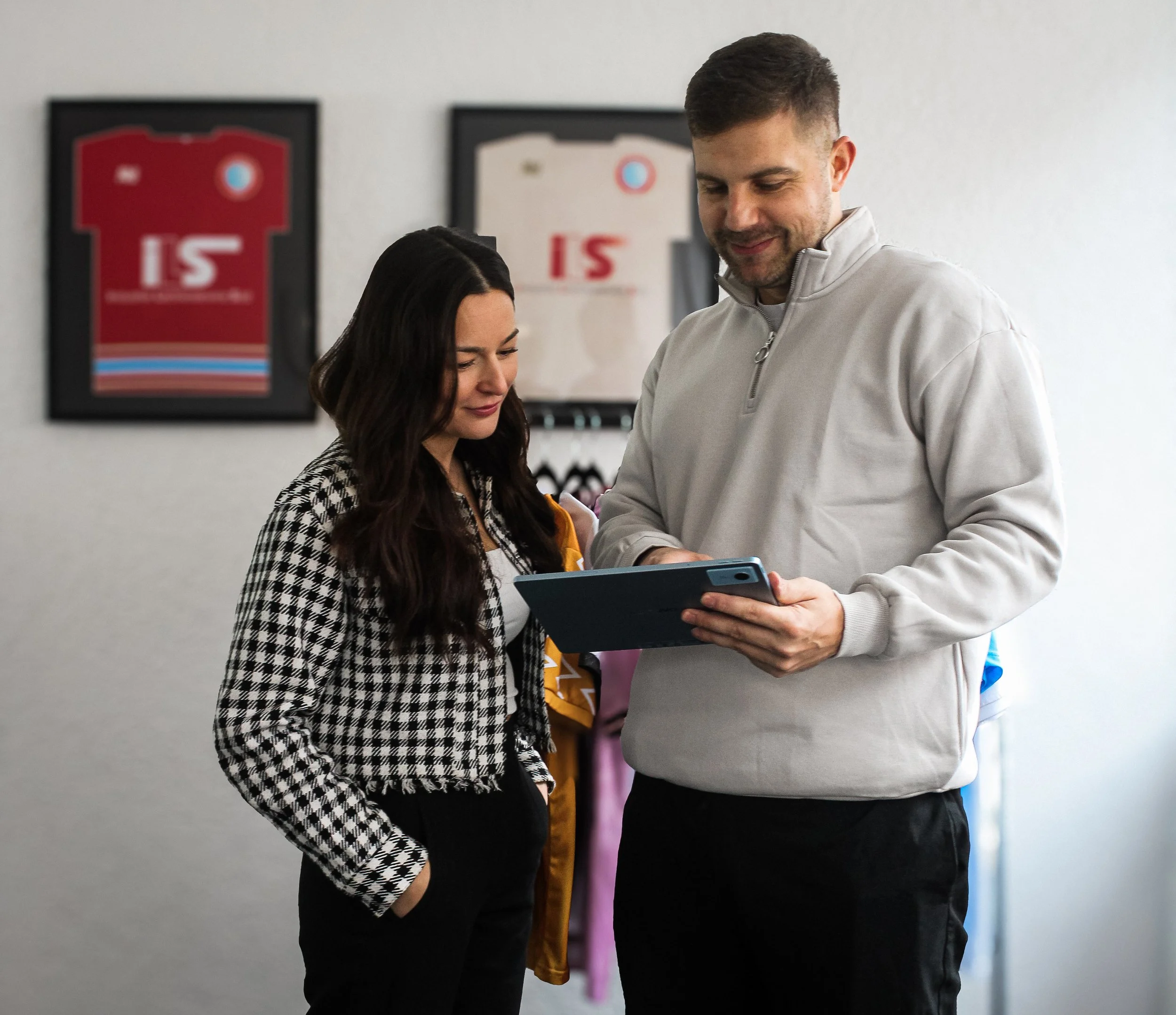 Ein Mann und eine Frau stehen zusammen und schauen auf ein Tablet. Im Hintergrund sind Trikots an der Wand. Der Mann trägt ein hellgraues Sweatshirt, die Frau trägt ein schwarz-weiß kariertes Jackett.