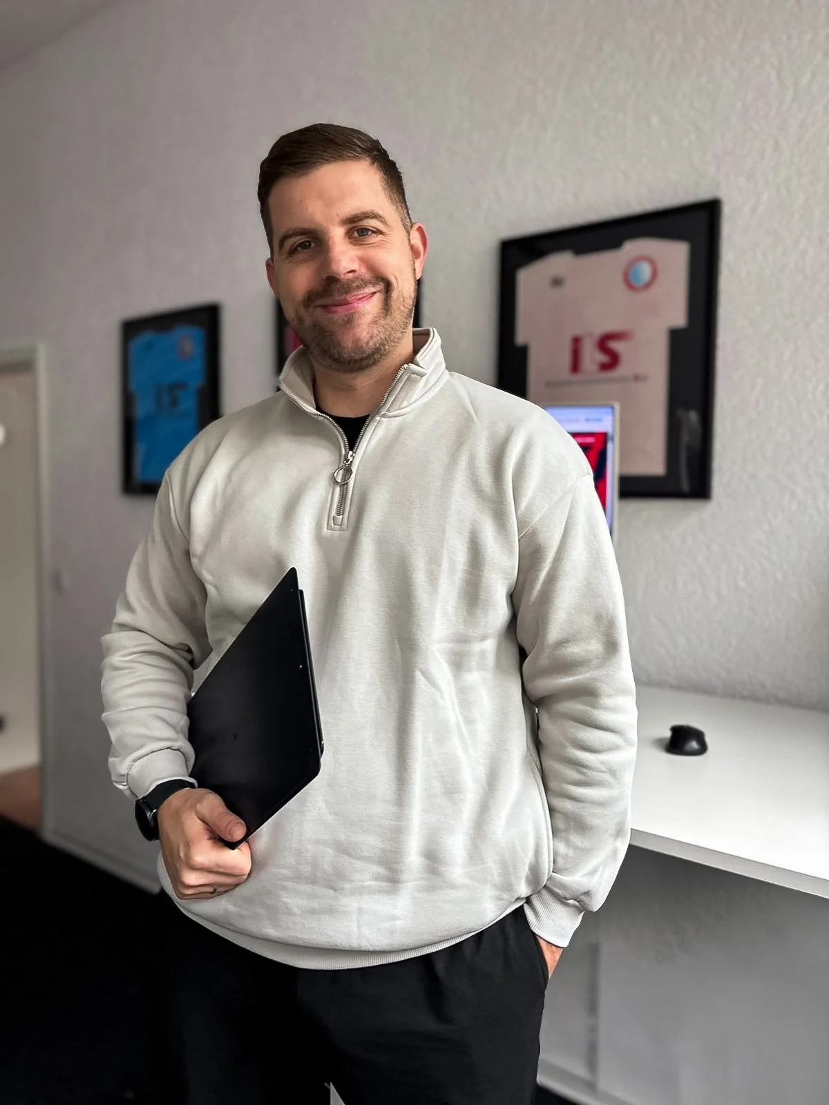 Ein Mann hält einen schwarzen Laptop in der Hand, trägt eine weiße Fleecejacke und lächelt, im Hintergrund sind Fußballtrikots und ein Computerbildschirm sichtbar.