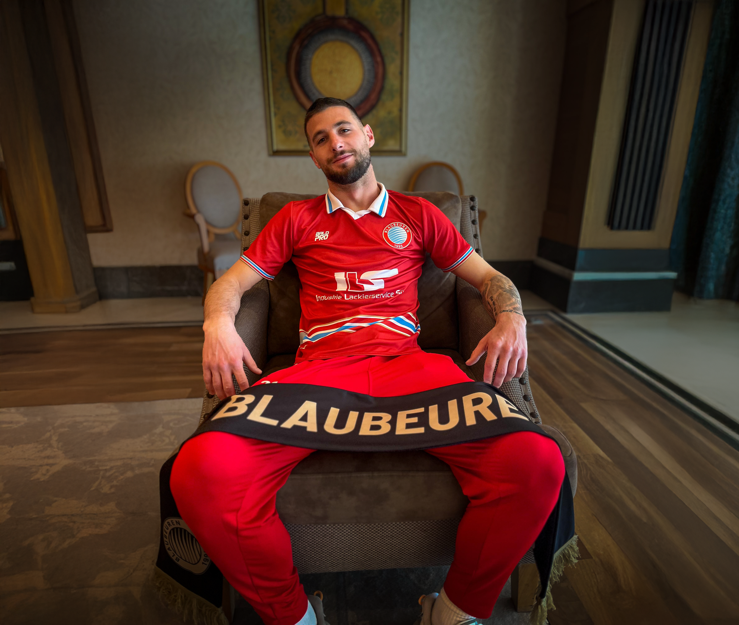 Mann in Rotem Fußballtrikot sitzt im Sessel im Interieur mit Holzboden, Wandbildern und Vorhängen.