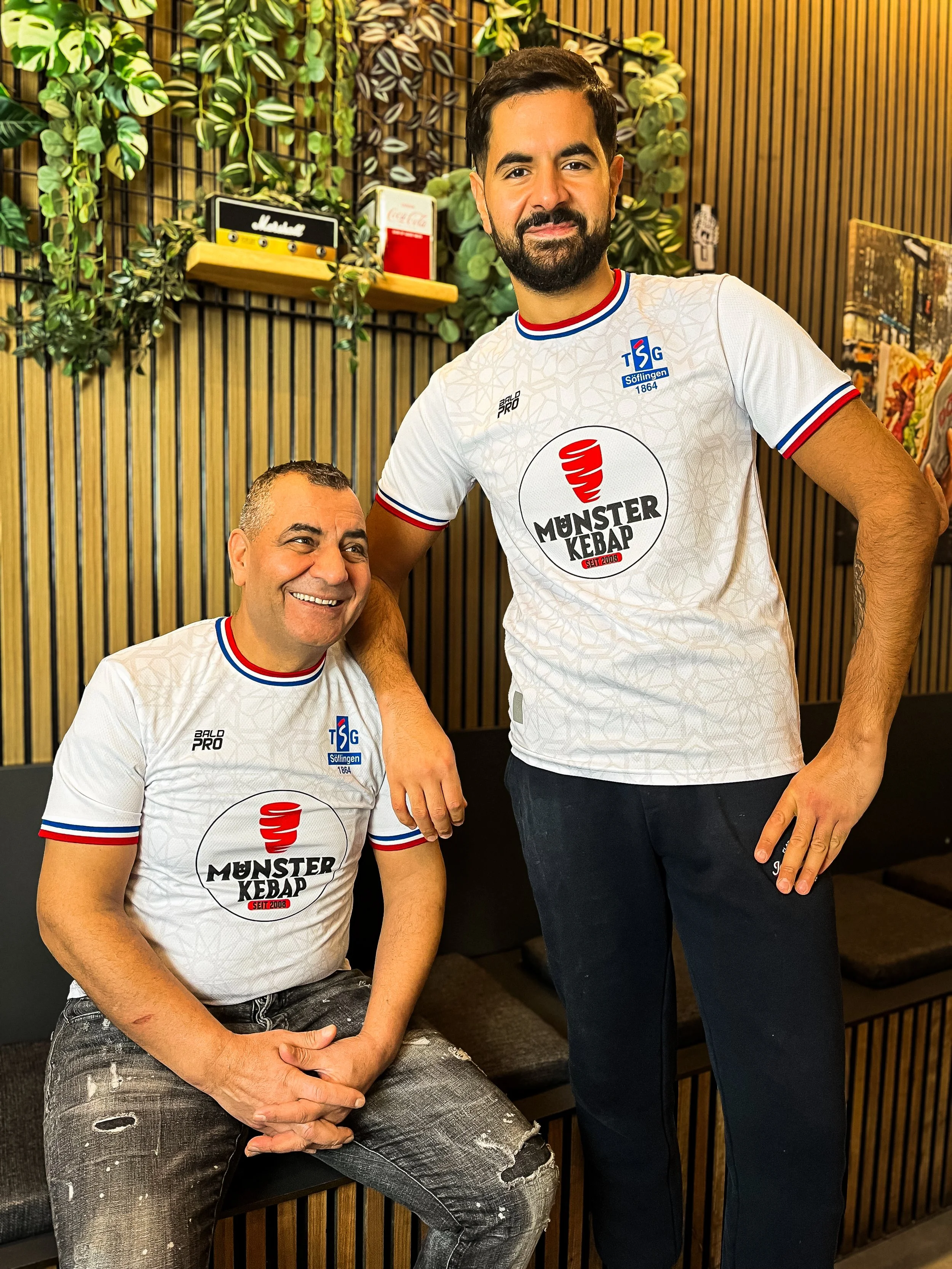 Zwei Männer in weißen Fußballtrikots mit dem Logo 'Münster Kebab' und dem Logo der TSG Söllingen posieren in einem Raum mit vertikalen Holzpaneelen und grünen Pflanzen im Hintergrund. Der sitzende Mann ist lächelnd, der stehende Mann legt seine Hand auf die Schulter des anderen.